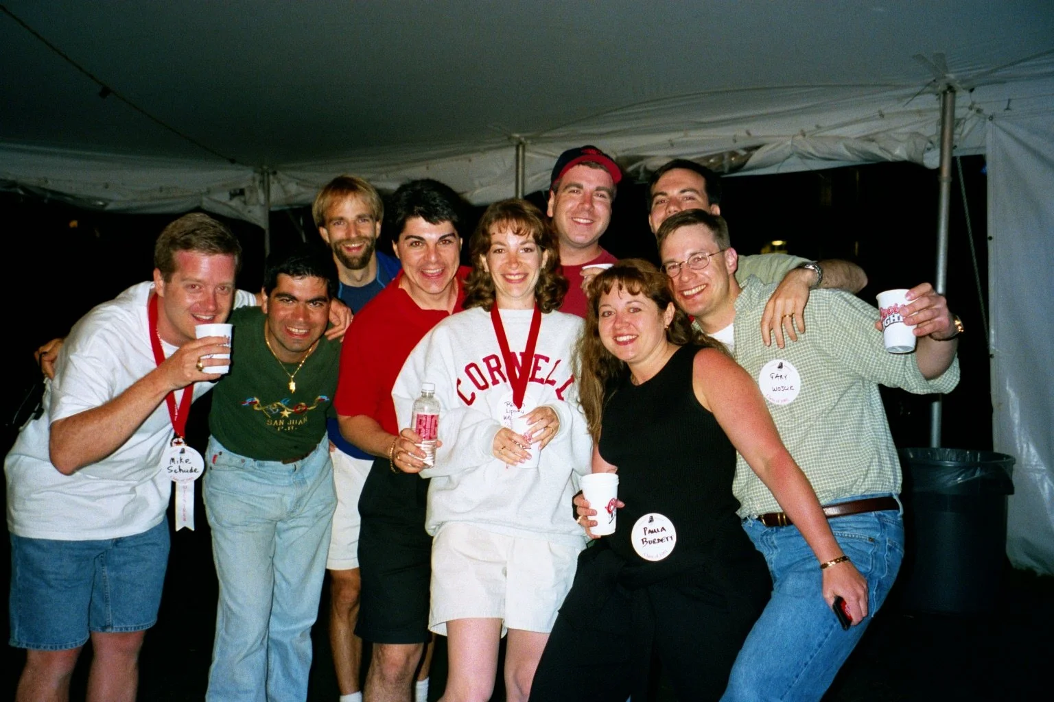Group of smiling people at an outdoor gathering, holding drinks, under a canopy, some wearing name tags, celebrating at night at the tent parties during Cornell Reunion weekend.