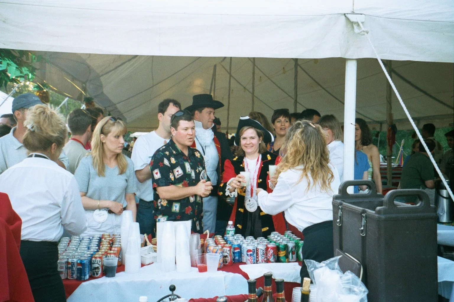 People gathered at a beverage stand under a large white tent during an outdoor event, some in costumes, with cans of soda on the table.