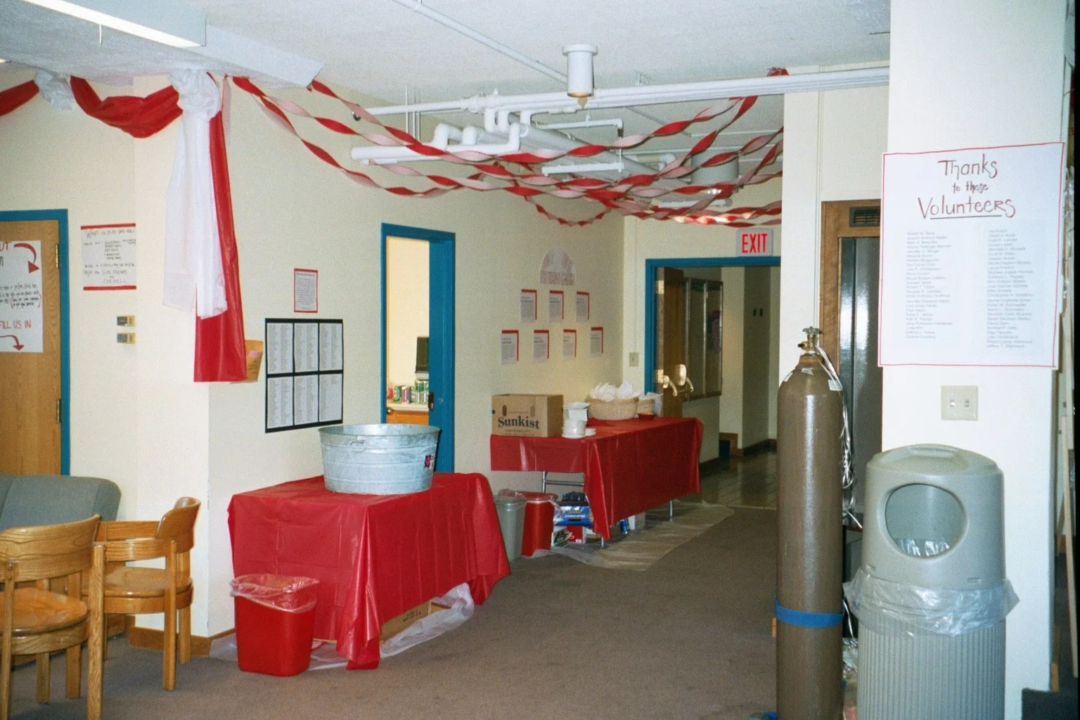 Indoor space decorated with red and white streamers hanging from the ceiling. Tables covered with red tablecloths are set up along the walls, holding various items. There's a large oxygen tank, a trash bin, chairs, and a wall with printed papers and 