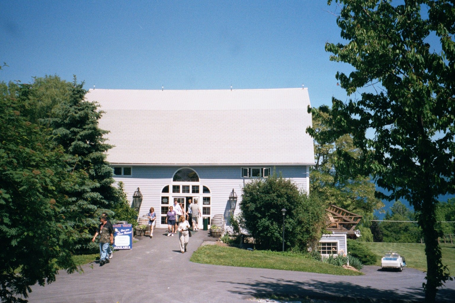 A white building with a large arched window entrance, surrounded by green trees and shrubs, with a small group of people walking towards it on a paved driveway and a vintage car parked nearby.