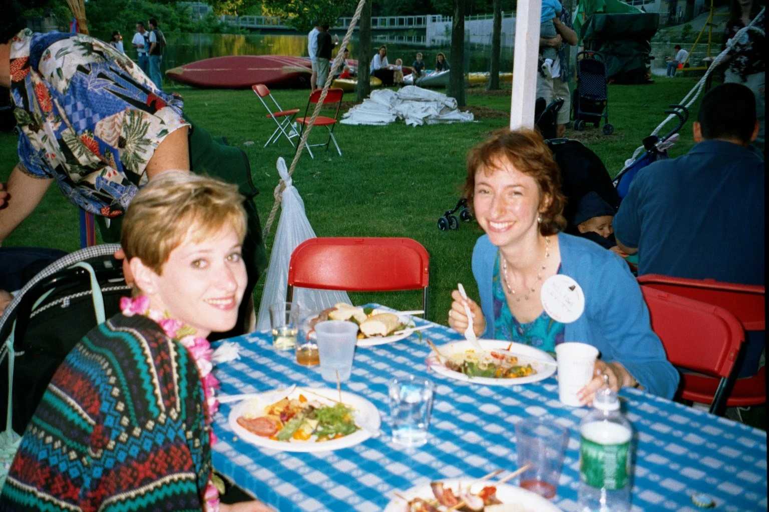 Two women sitting at a picnic table with blue and white checkered tablecloth, enjoying food at an outdoor gathering by a lake. One woman has short blonde hair and is wearing a colorful patterned sweater and pink lei, while the other woman has short b