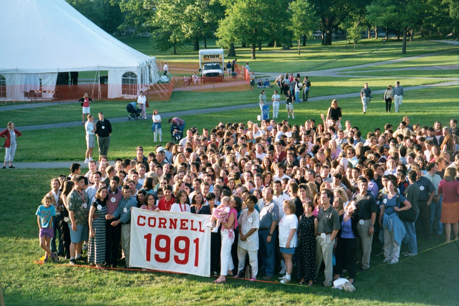 A large group of people gathered outdoors on a grassy area, holding a banner that reads 'CORNELL 1991'. There are tents and trees in the background, with some individuals standing and walking around.