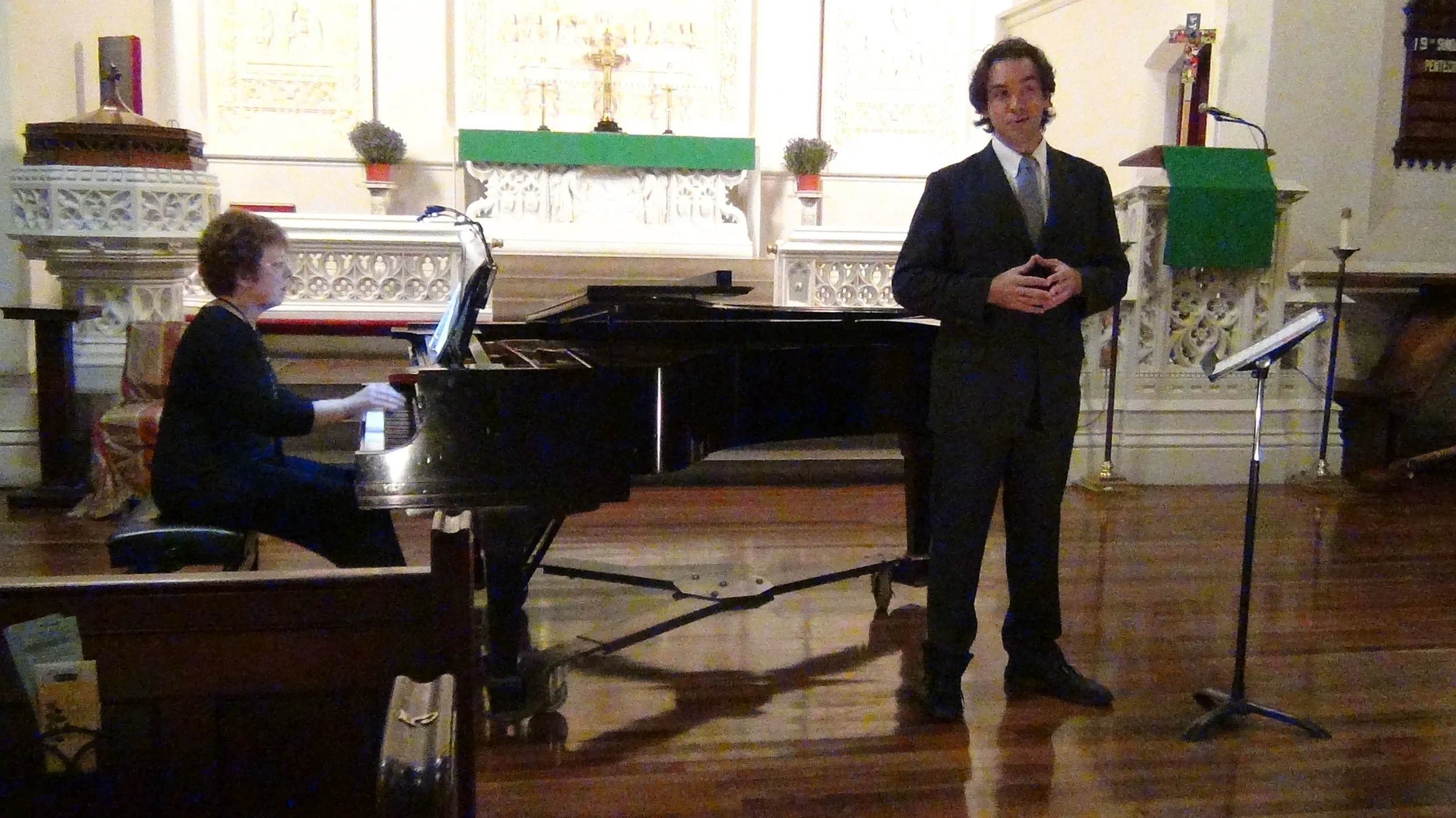 Woman playing grand piano, man in suit standing with hands clasped, in a church or chapel setting with religious decorations and candles.