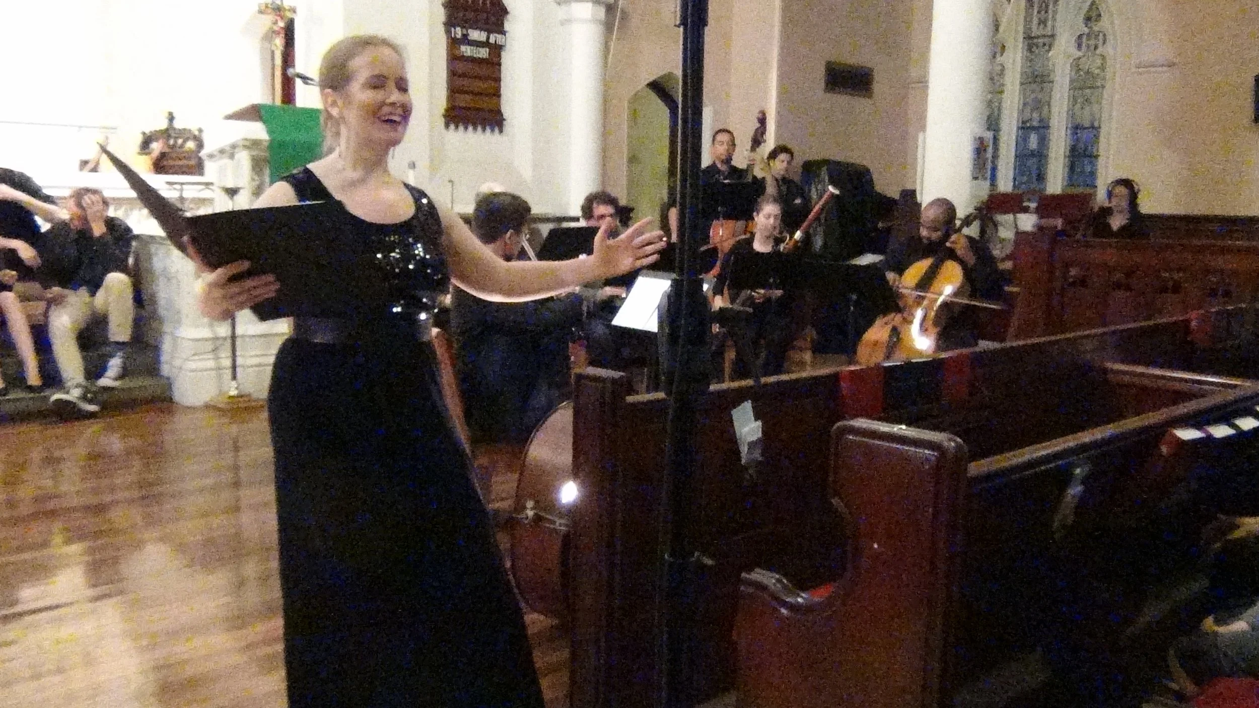 A woman in a black dress is singing or speaking with a smile in a church or concert hall, with musicians playing string instruments in the background.