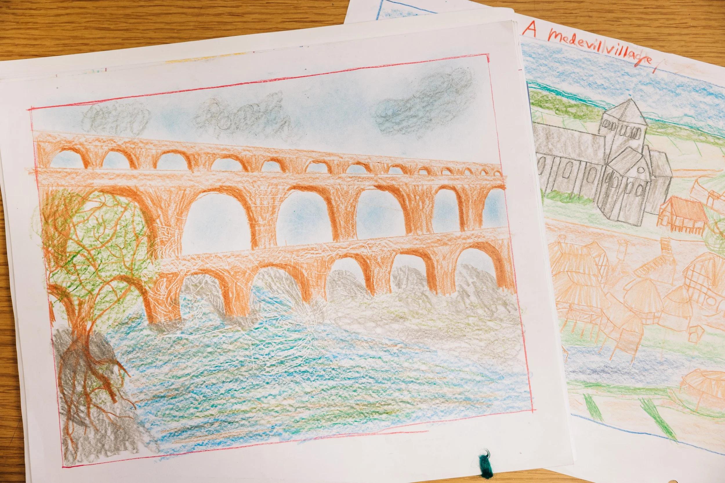 Colorful hand-drawn pictures of a bridge over a river and a church in a village on white paper on a wooden surface.