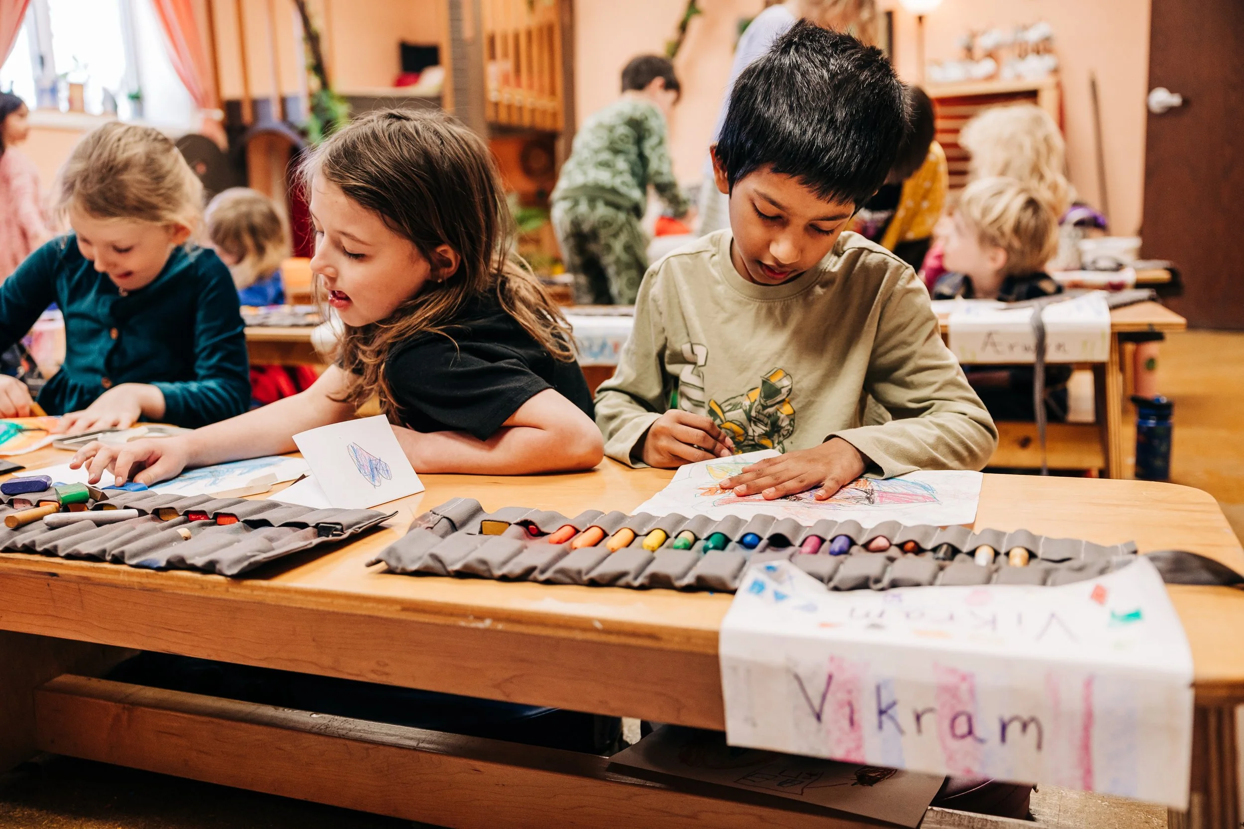 Children sitting at a wooden table, engaged in drawing and coloring activities with crayons and markers, in a classroom setting.