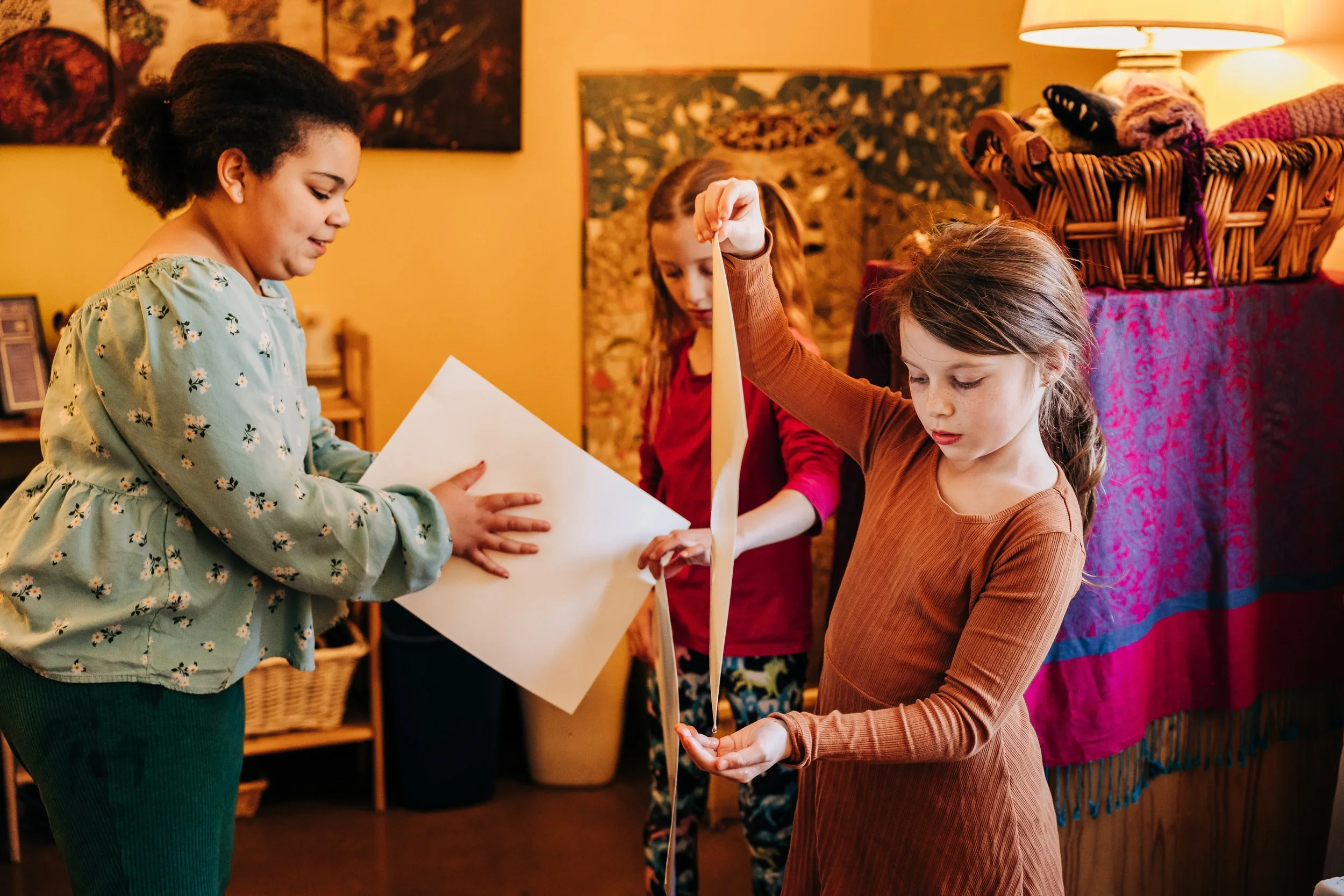 Three young girls in a cozy room with warm yellow lighting, one holding a large blank piece of paper and another holding a long strip of paper, possibly making a craft or participating in an activity.