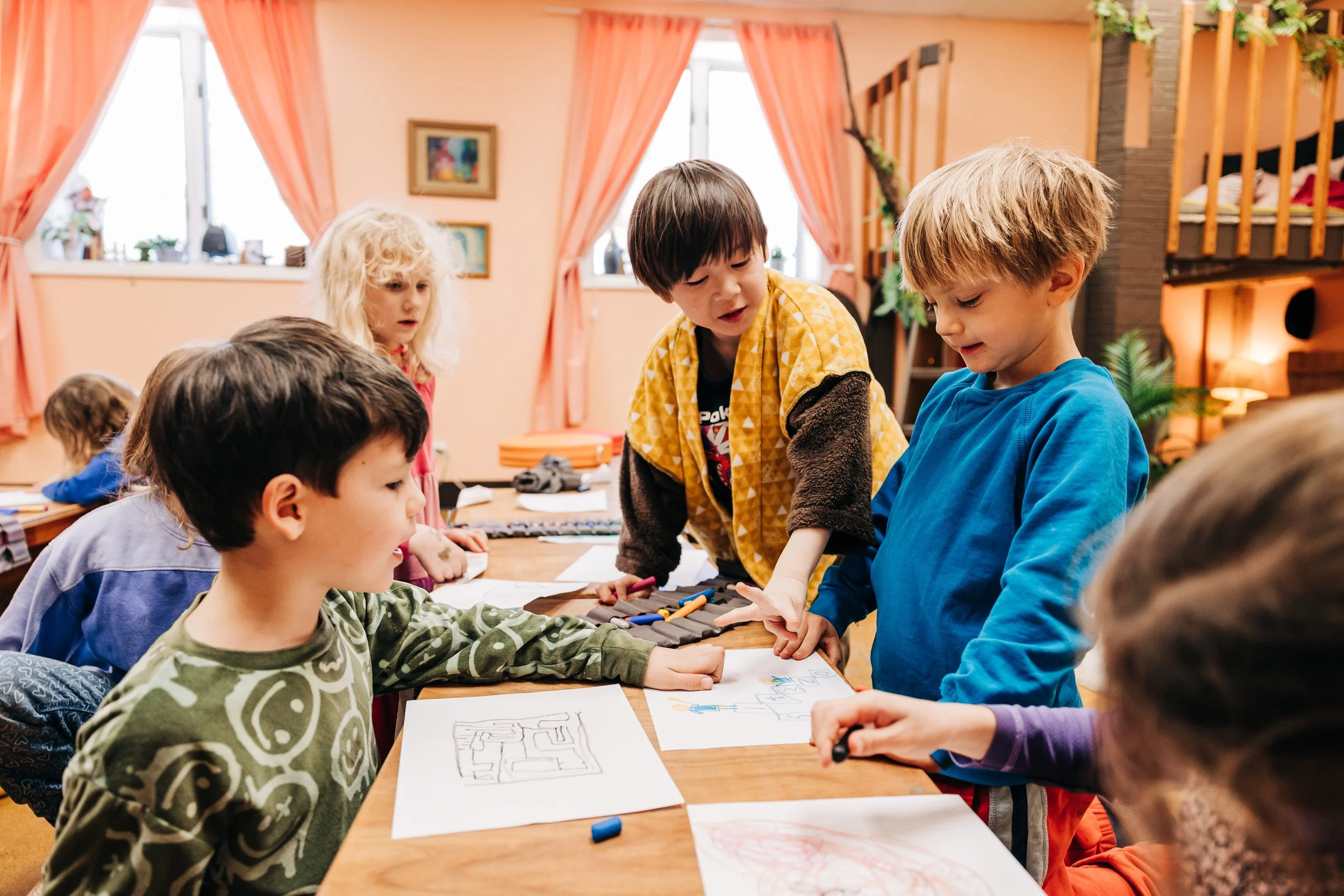 Children gathered around a table drawing and discussing artwork in a cozy, well-lit room with peach-colored walls, pink curtains, and decorative elements.