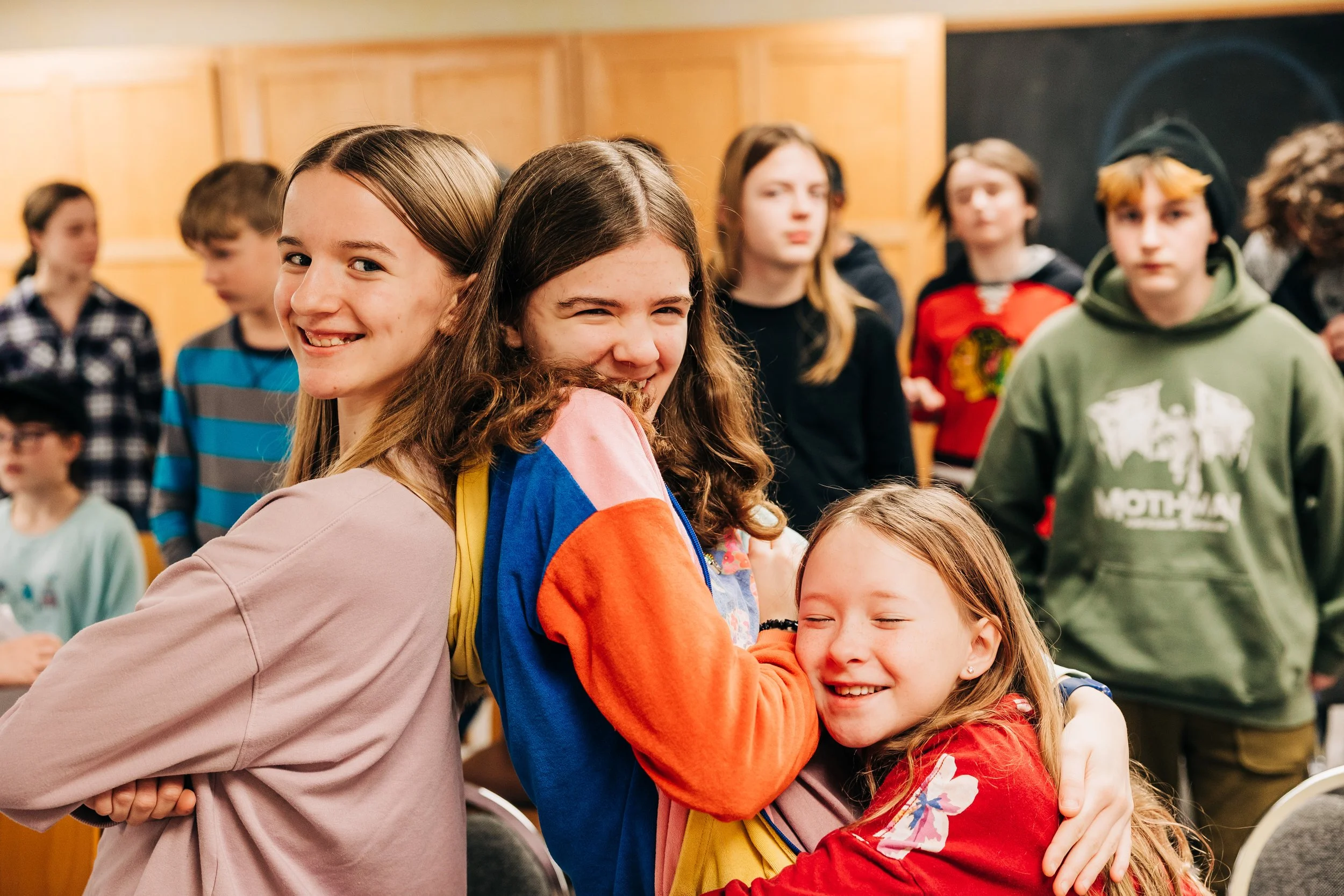 Three smiling girls hugging in a group of children in a room with wood-paneled walls.