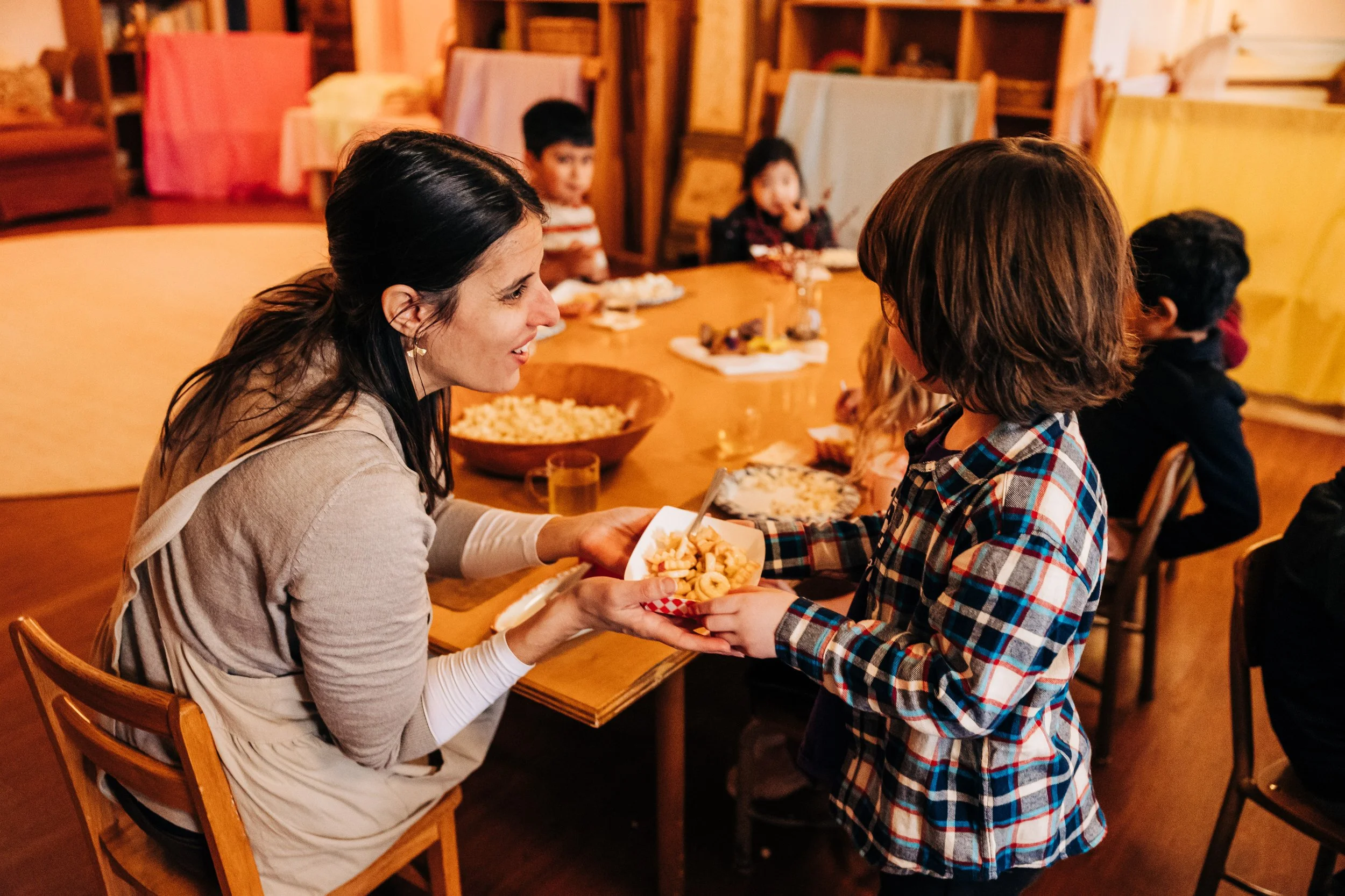 Woman is handing a plate of food to a young boy in an early childhood classroom with other kids seated at a table in the background.