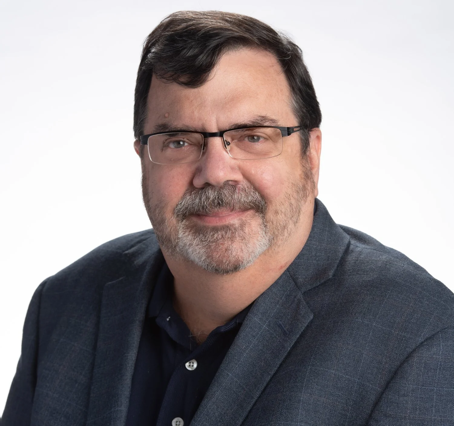 Professional headshot of a middle-aged man with dark hair, glasses, and a beard, wearing a dark suit jacket and a navy shirt, smiling against a white background.