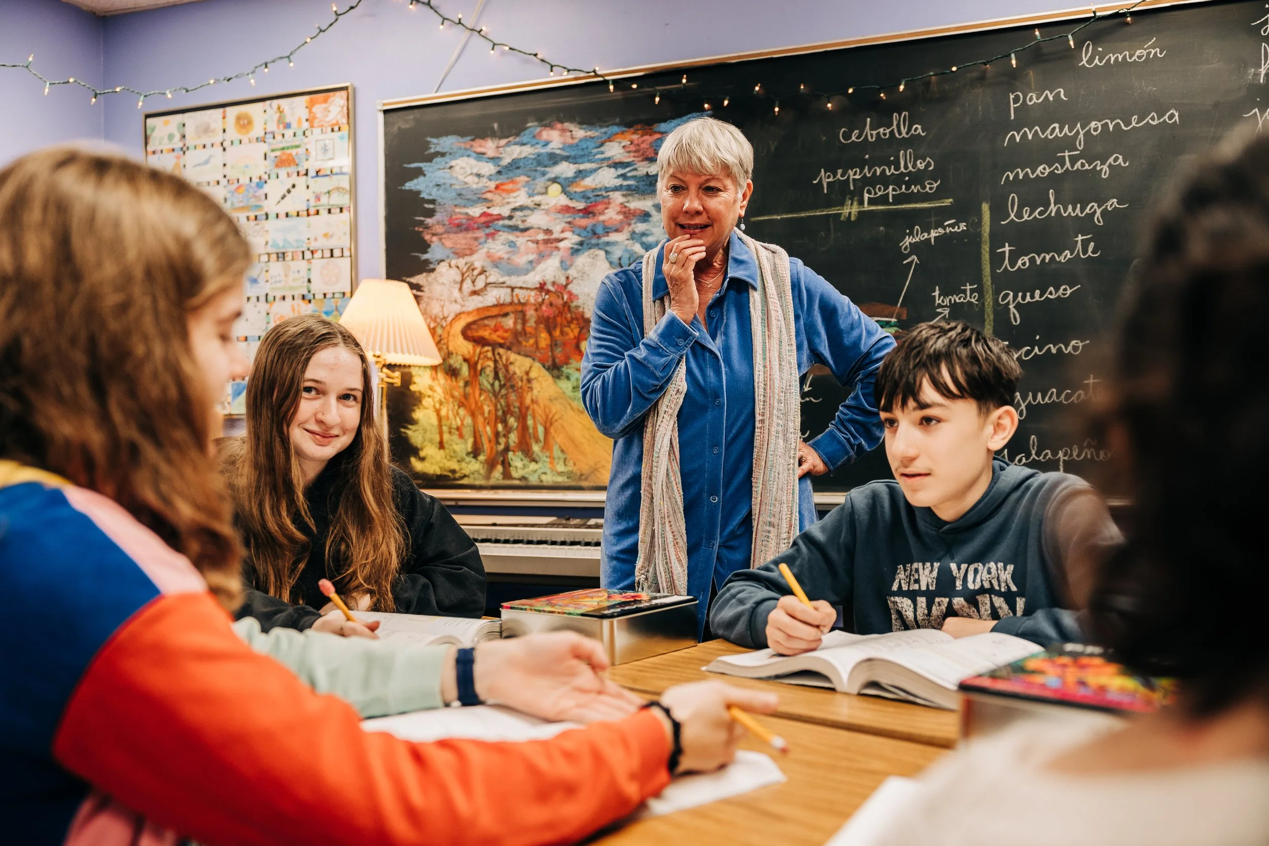 A teacher standing at the front of a classroom with students sitting at desks around a table, blackboard with writing behind her, and a colorful mural on the wall.