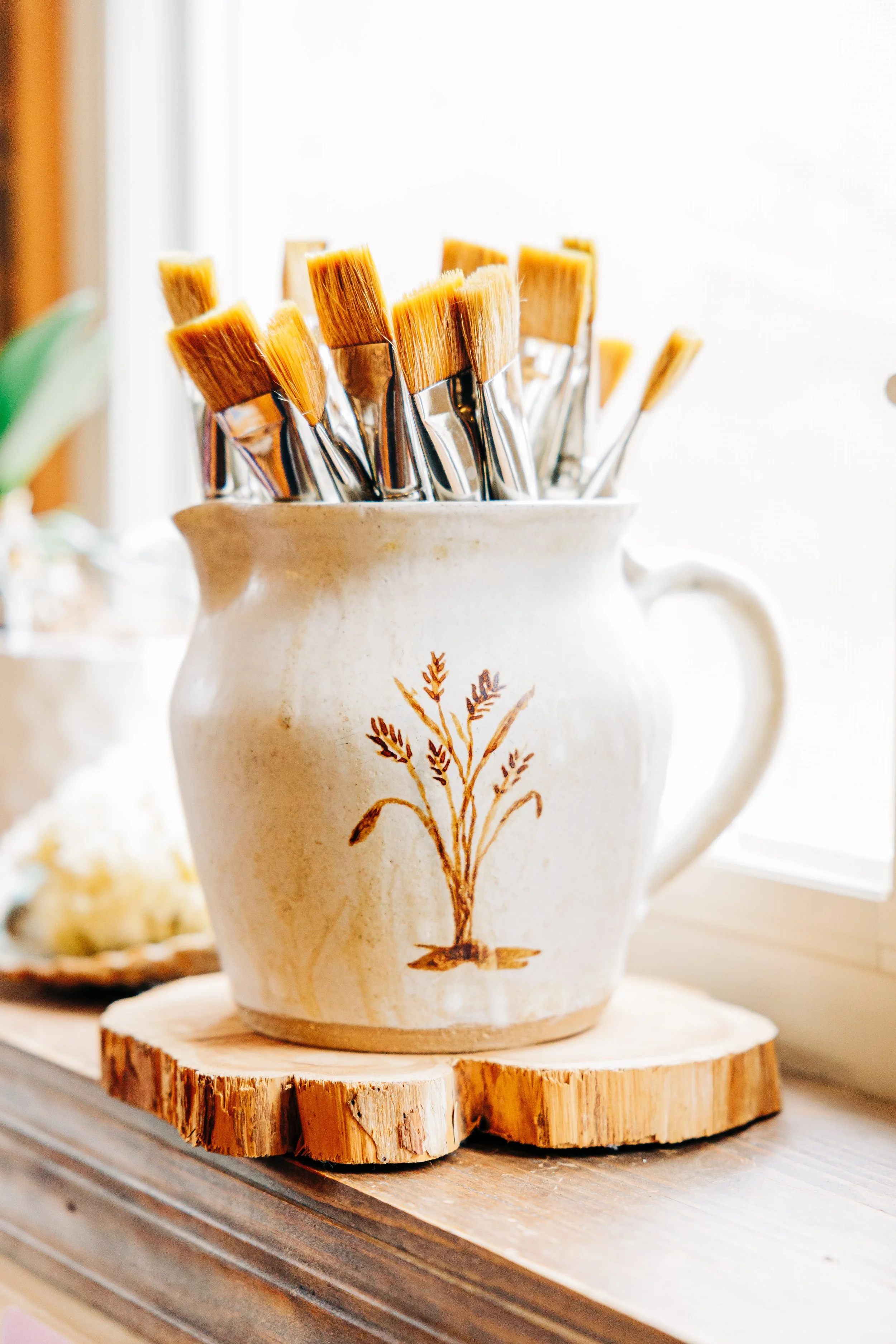 Ceramic pitcher with a wheat design holding paintbrushes of various sizes near a window.