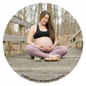 Une femme enceinte assise en tailleur sur une plateforme en bois dans la nature, tenant son ventre avec les mains, portant un débardeur noir et un pantalon violet, souriant.