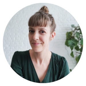 Portrait d'une femme souriante avec une coiffure en chignon, portant un haut vert, devant un mur blanc avec une plante en arrière-plan.