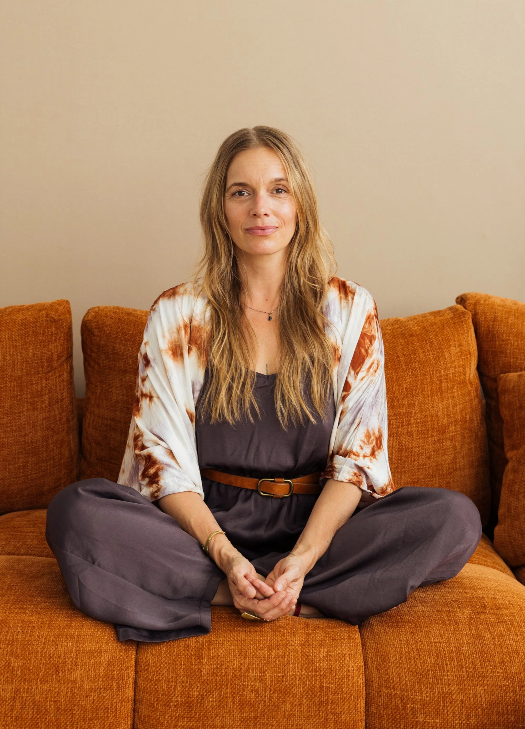 Une femme aux cheveux longs et blonds, assise en position de croissant sur un canapé orange, portant une robe grise avec une ceinture marron, un kimono blanc avec des motifs marron, et regardant directement la caméra.