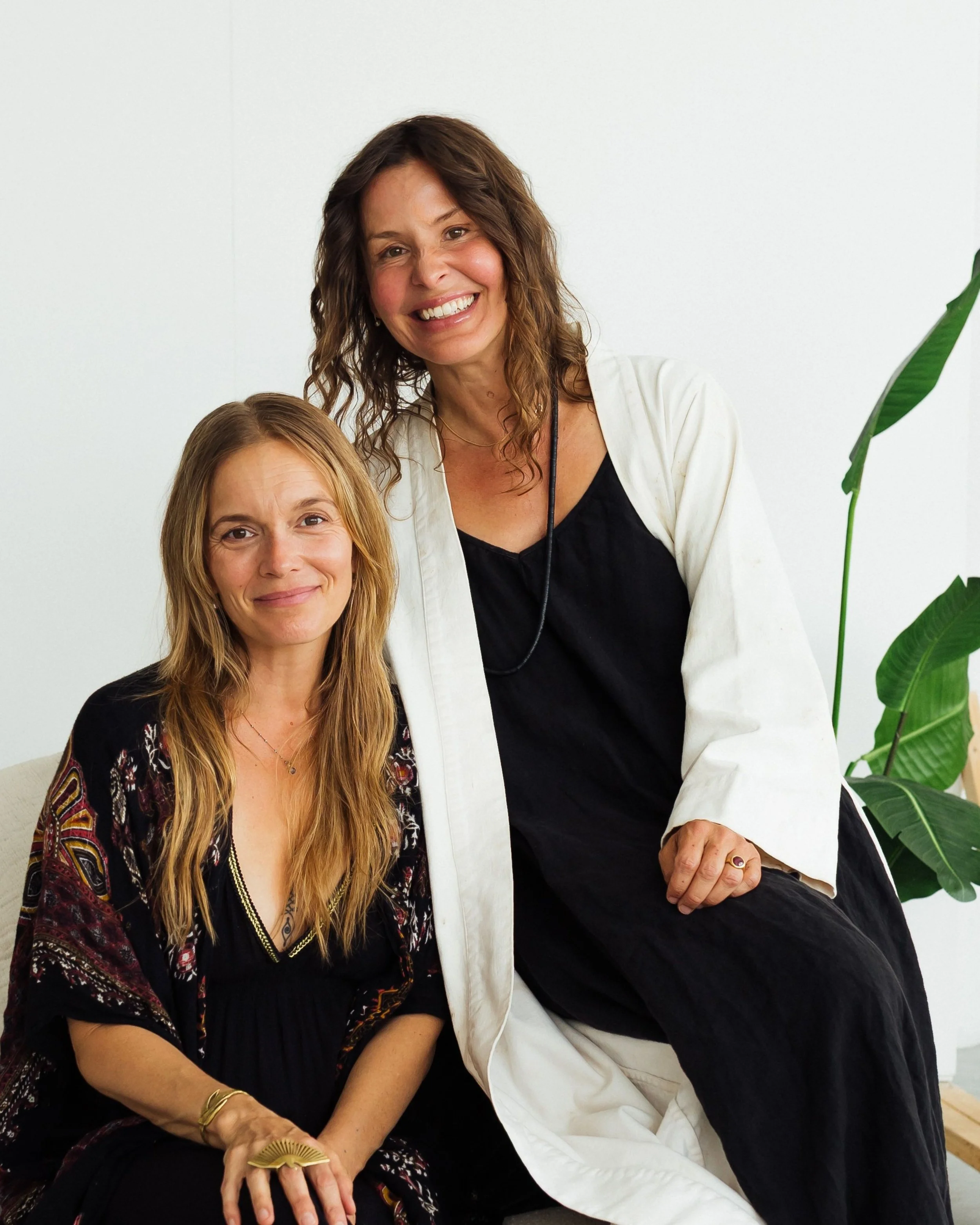 Deux femmes, une assise et une debout, souriant, dans un intérieur avec un plante verte en arrière-plan.