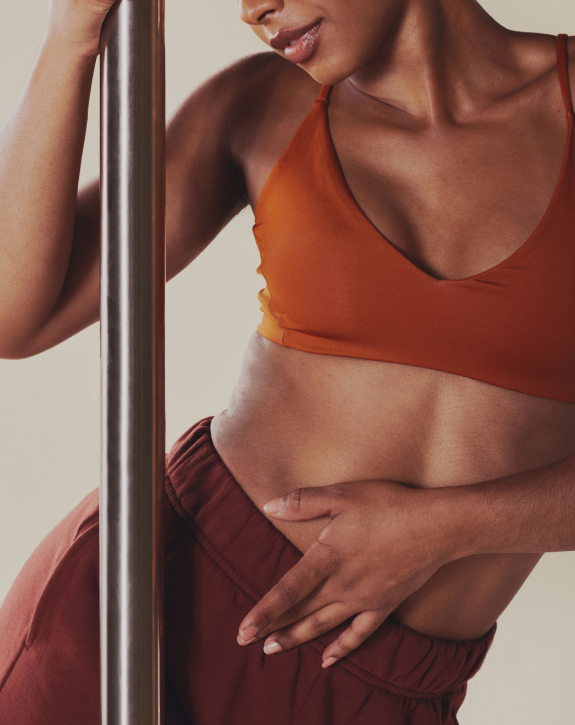 Close-up of a woman in fitness attire holding a pole, with her hand on her stomach, wearing an orange sports bra and maroon sweatpants.