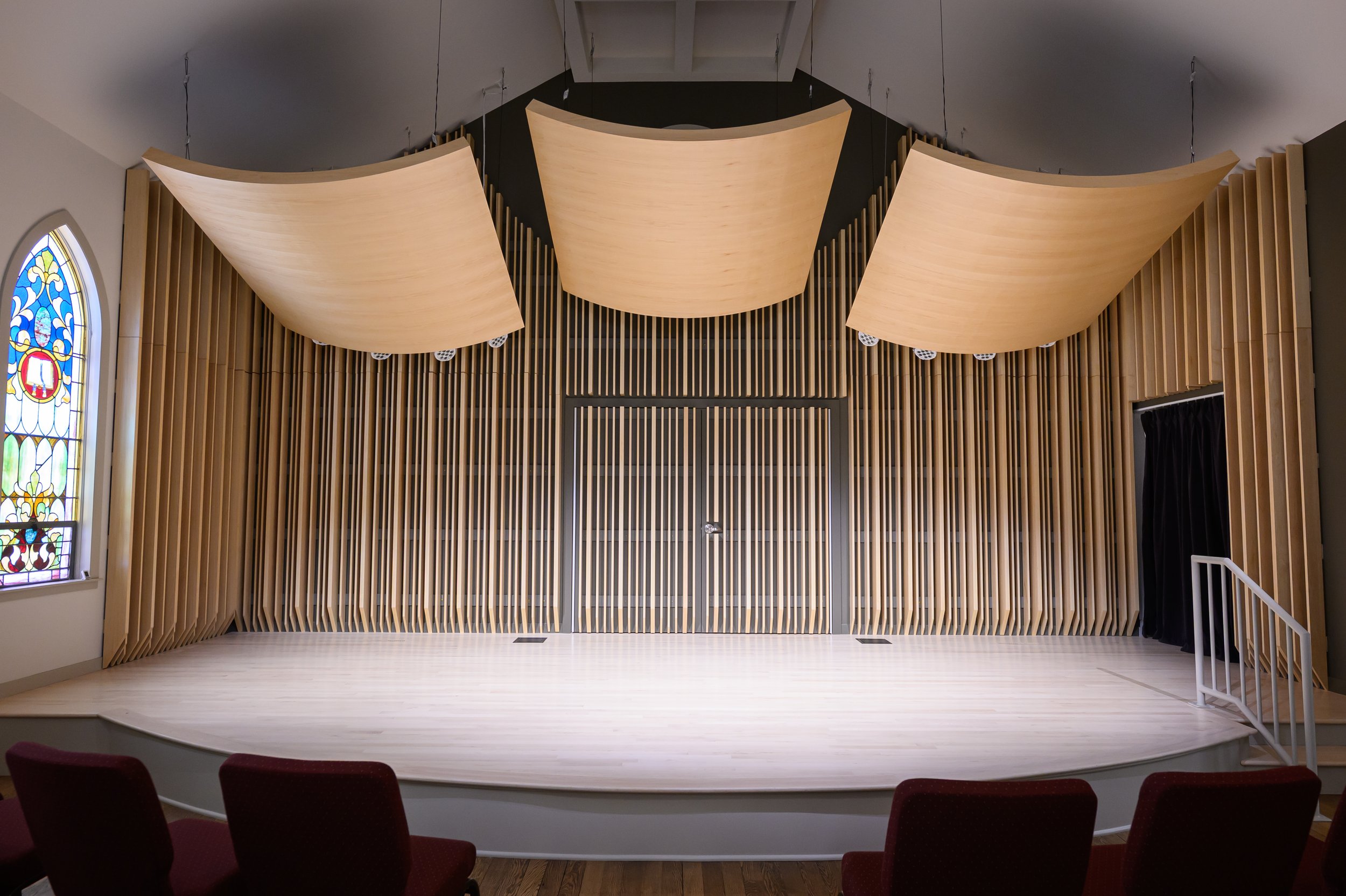 Empty stage with wooden slats and large curved wooden sound clouds overhead, stained glass window on the left, black curtain on the right, burgundy chairs in the foreground.