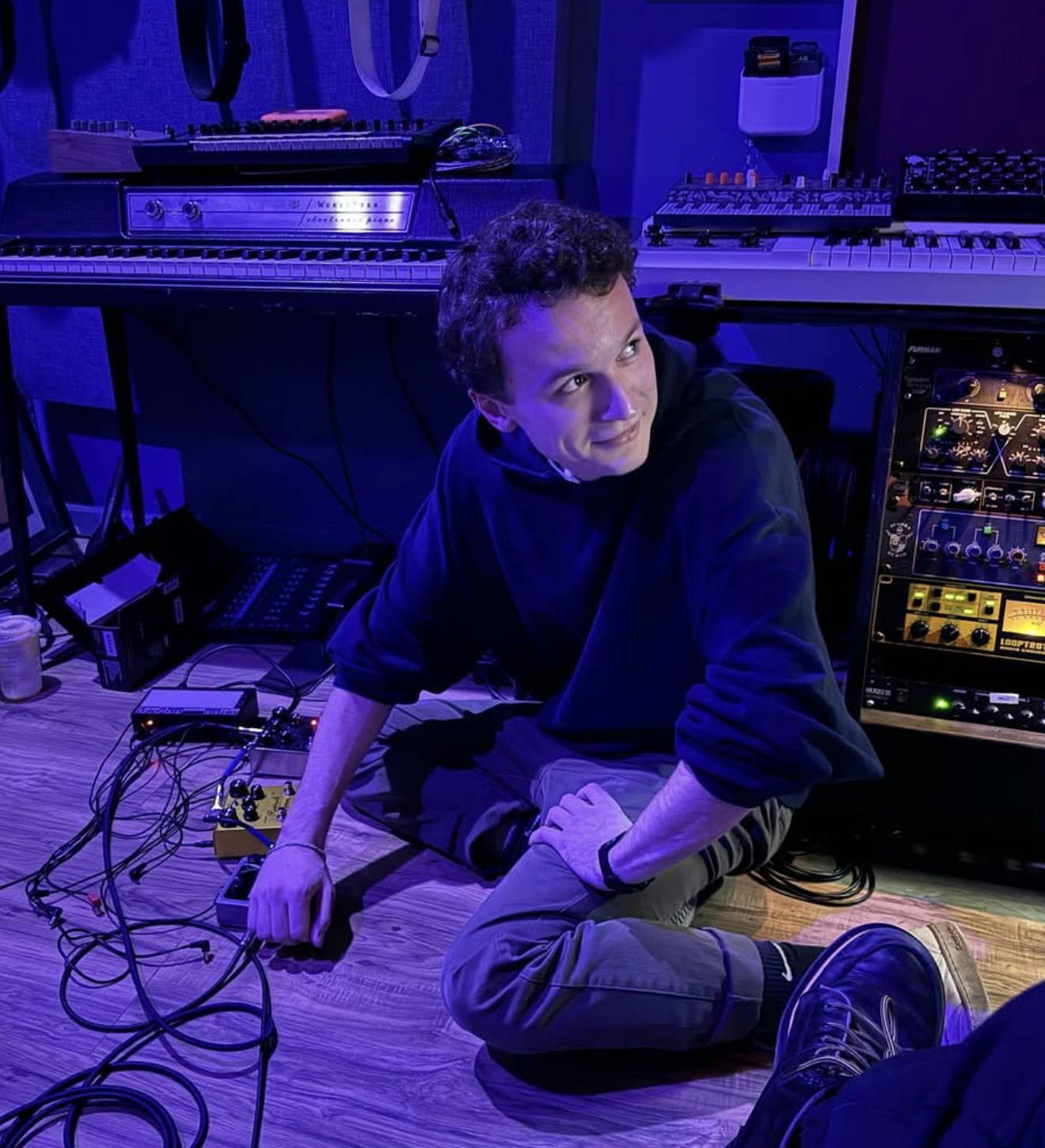 A person with curly hair in a black hoodie and khaki pants sitting on a wooden floor in a music studio surrounded by electronic music equipment, including a keyboard, synthesizer, and various cables.