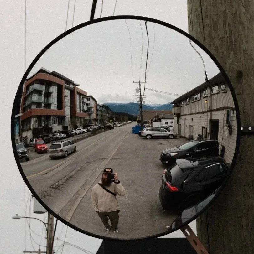 Reflection of a person taking a photo in a circular mirror, capturing a street scene with parked cars, buildings, and mountains in the background.