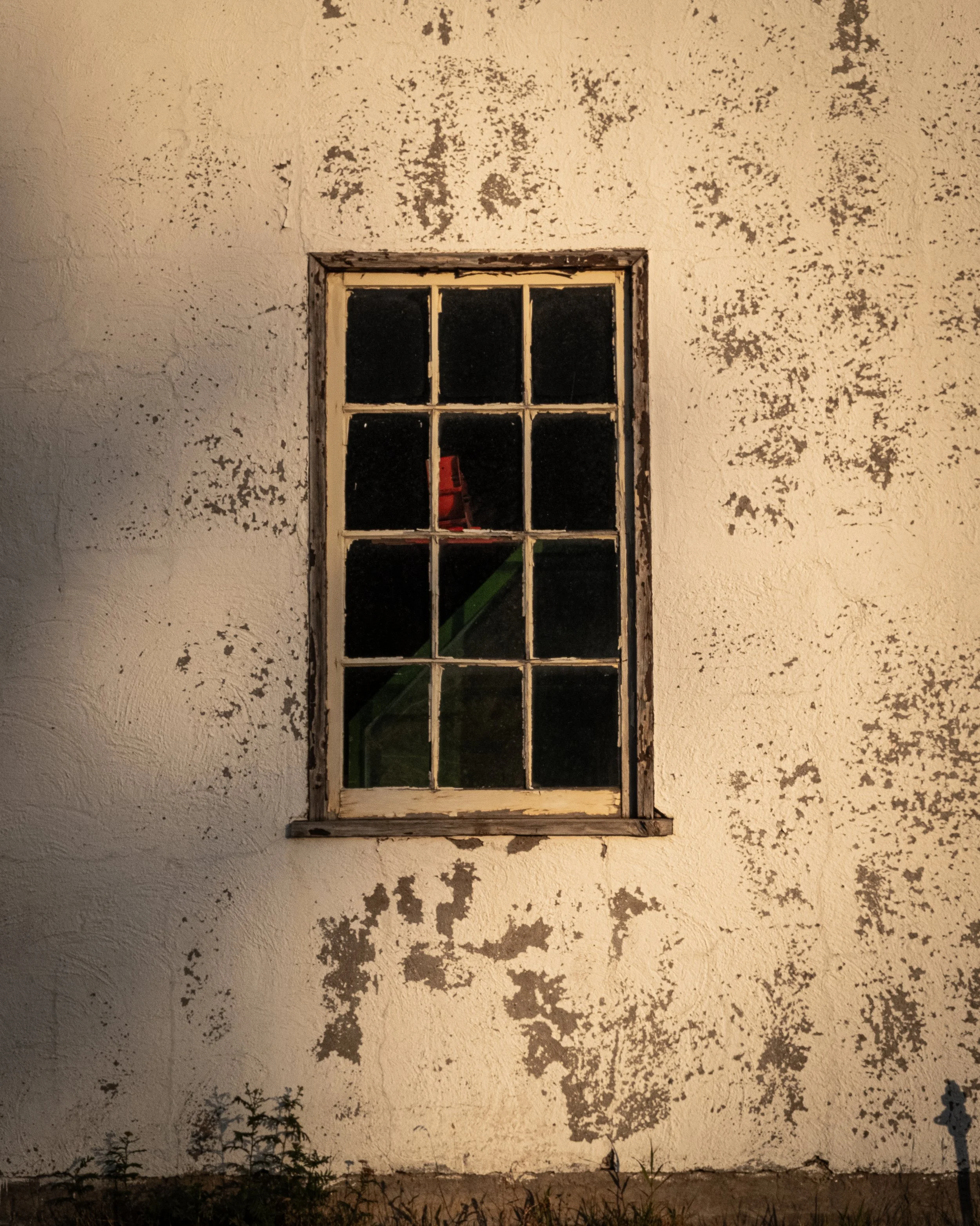 A weathered, white stucco wall with peeling paint and an old window with multiple panes. The window reveals a red chair inside and some green structure behind it.