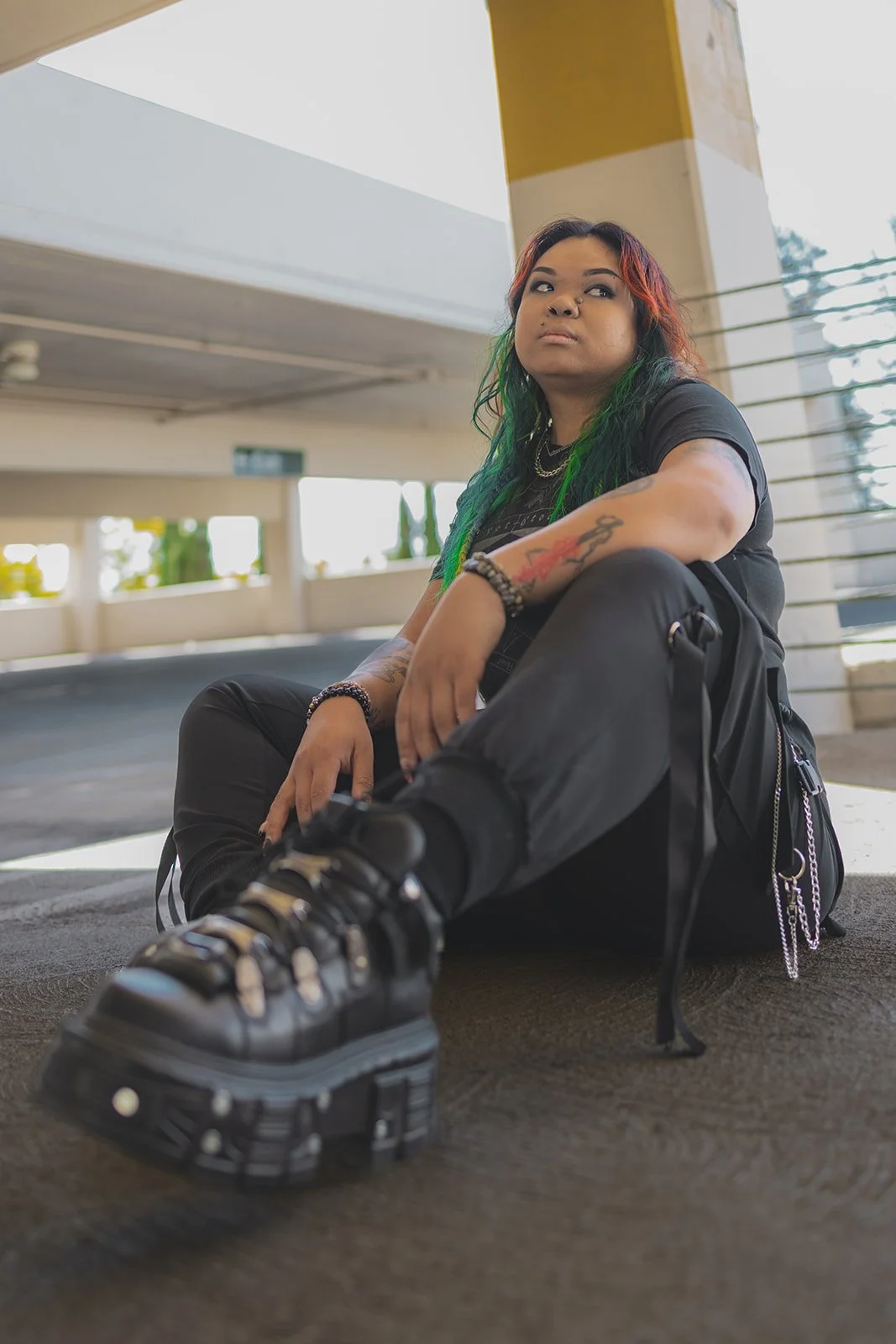 Chetto Thach sitting in a parking garage in Midtown Sacrametno wearing black boots with rainbow green and orange hair.