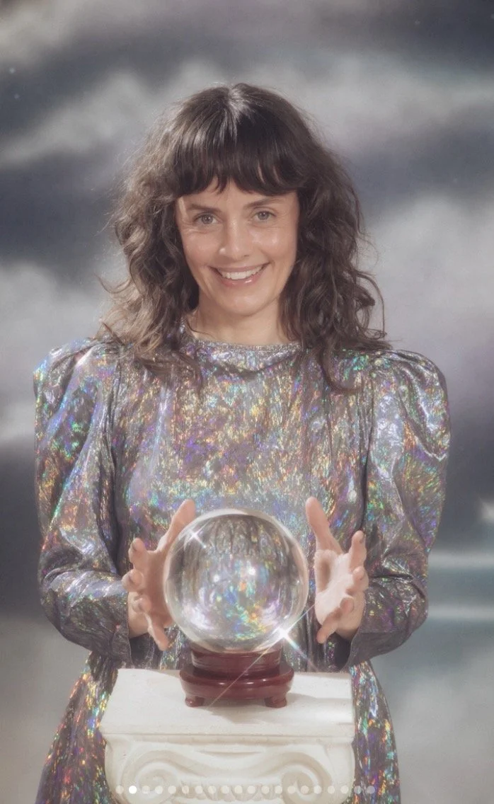A woman with shoulder-length brown curly hair wearing a shiny, iridescent dress, standing behind a crystal ball on a white column. She is smiling and holding the crystal ball with both hands. The background is gray and purple with misty clouds.