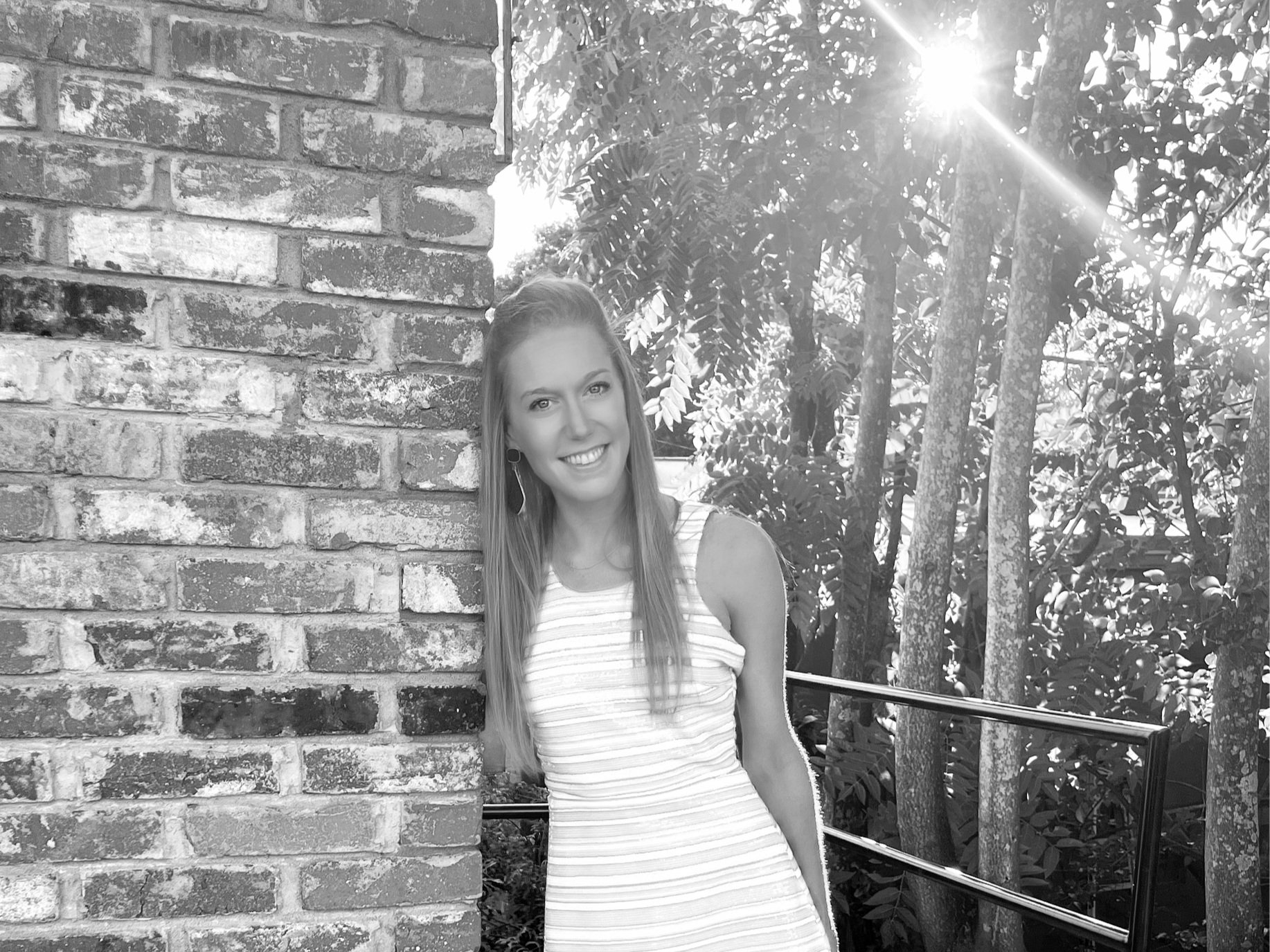 A woman with long hair and earrings smiling, standing next to a brick wall and a metal railing outdoors, with sunlight and trees in the background.