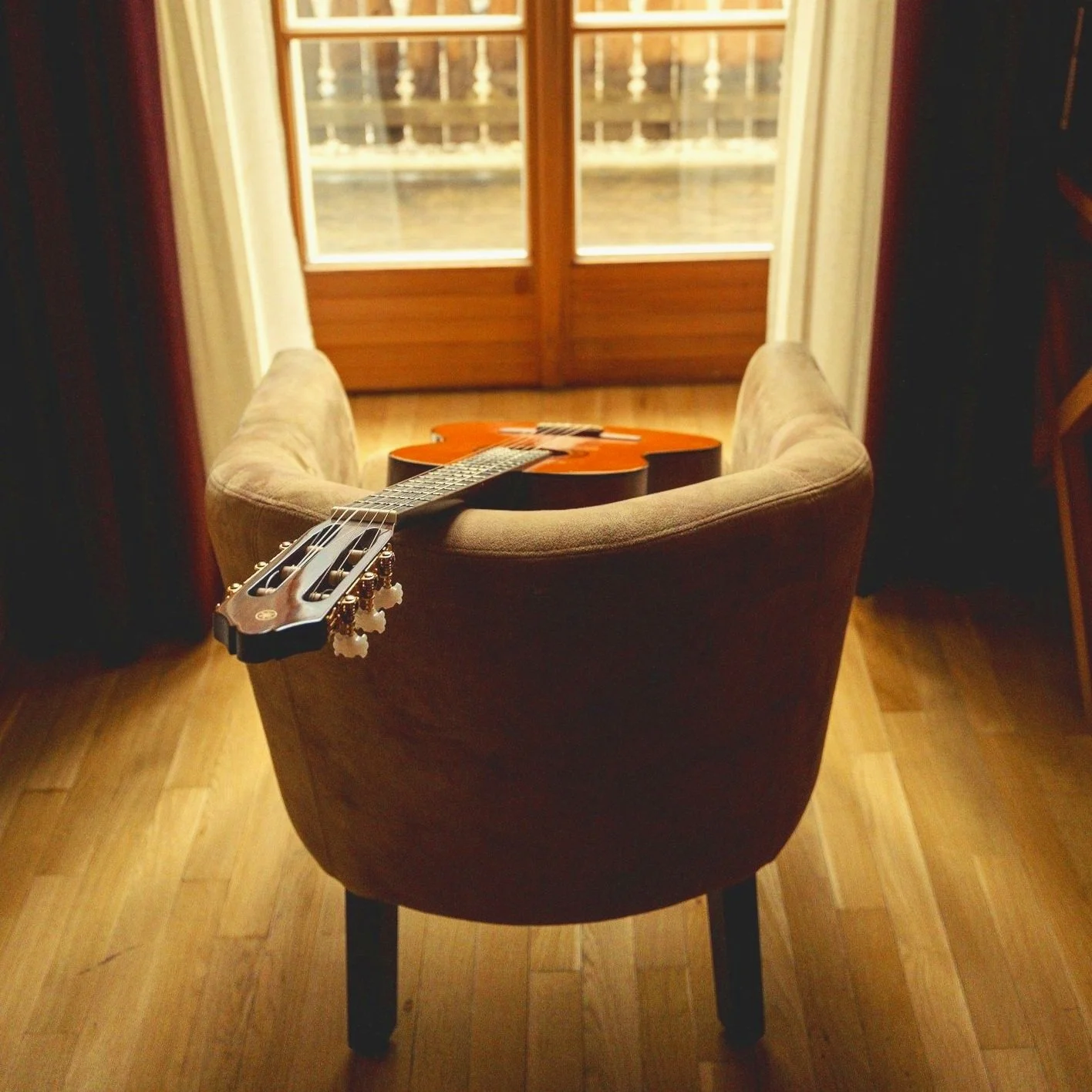 Acoustic guitar resting on a rounded, brown upholstered armchair in front of a wooden door with glass panes, overlooking a balcony.
