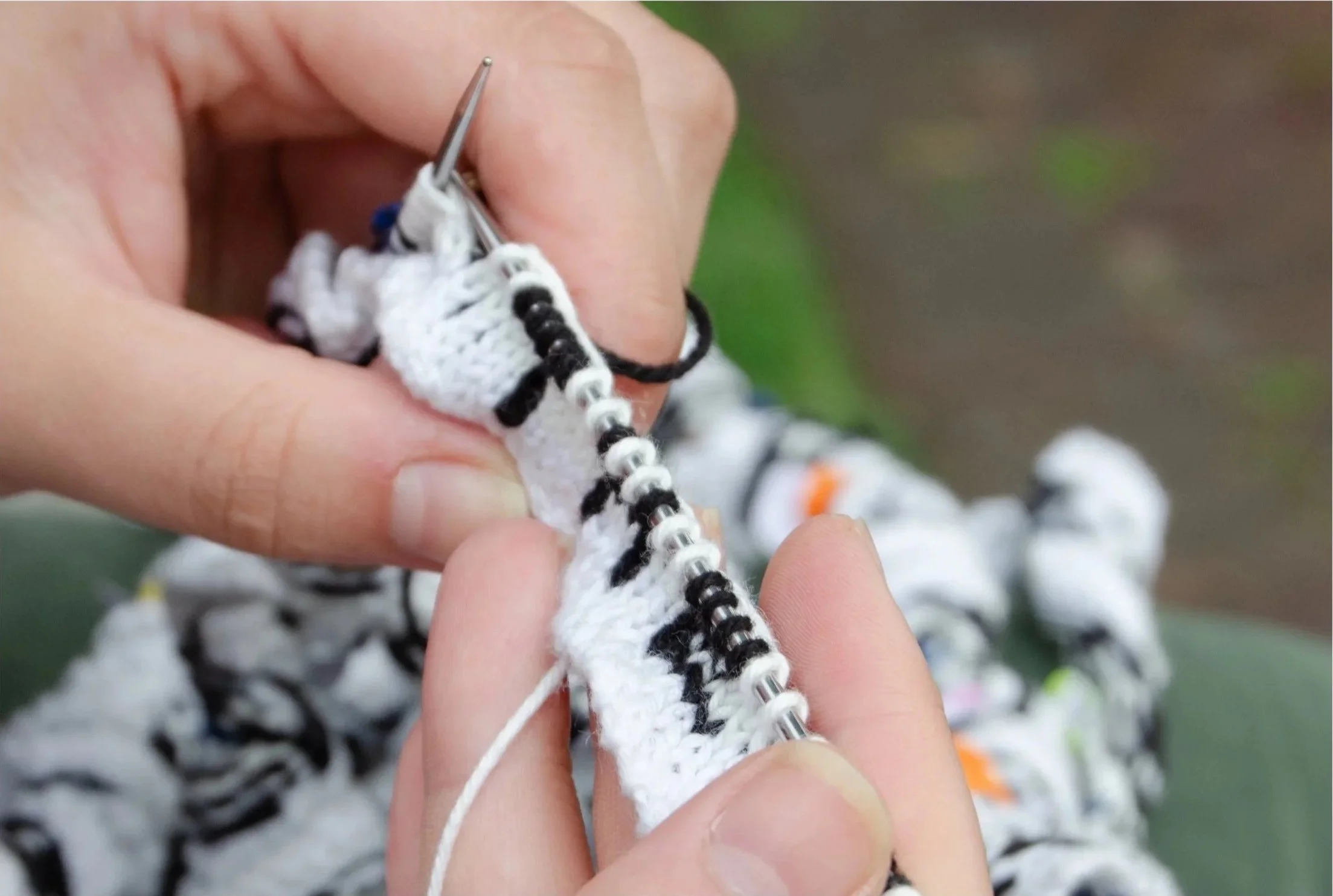 Photograph of artist's hands knitting the ribbon, with a portion showing the word "THE" on the needles.