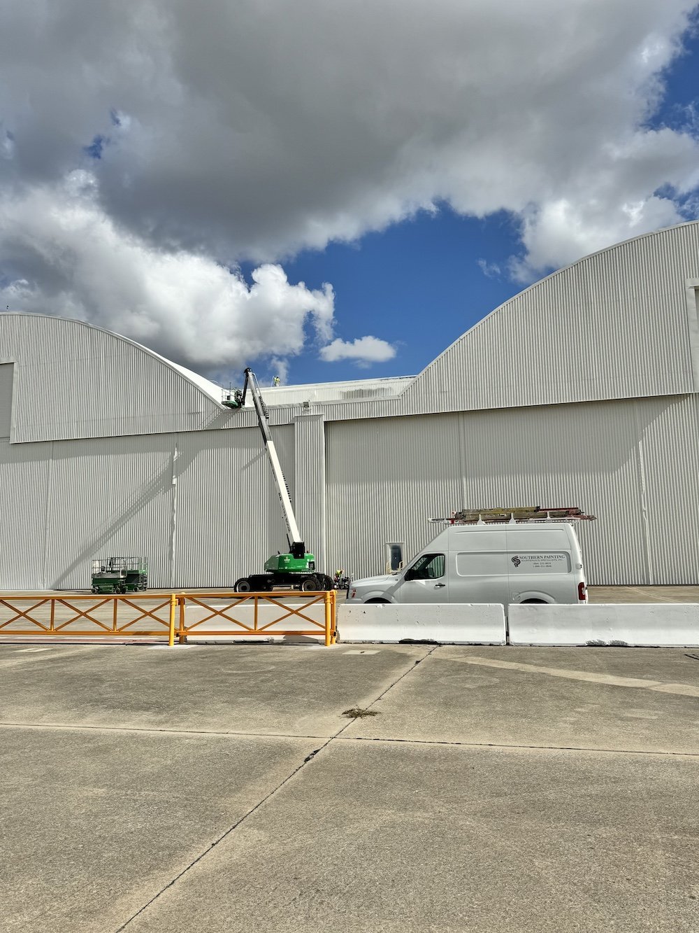 Workers on elevated lifts making repairs on the white exterior of a large industrial building under a partly cloudy sky.