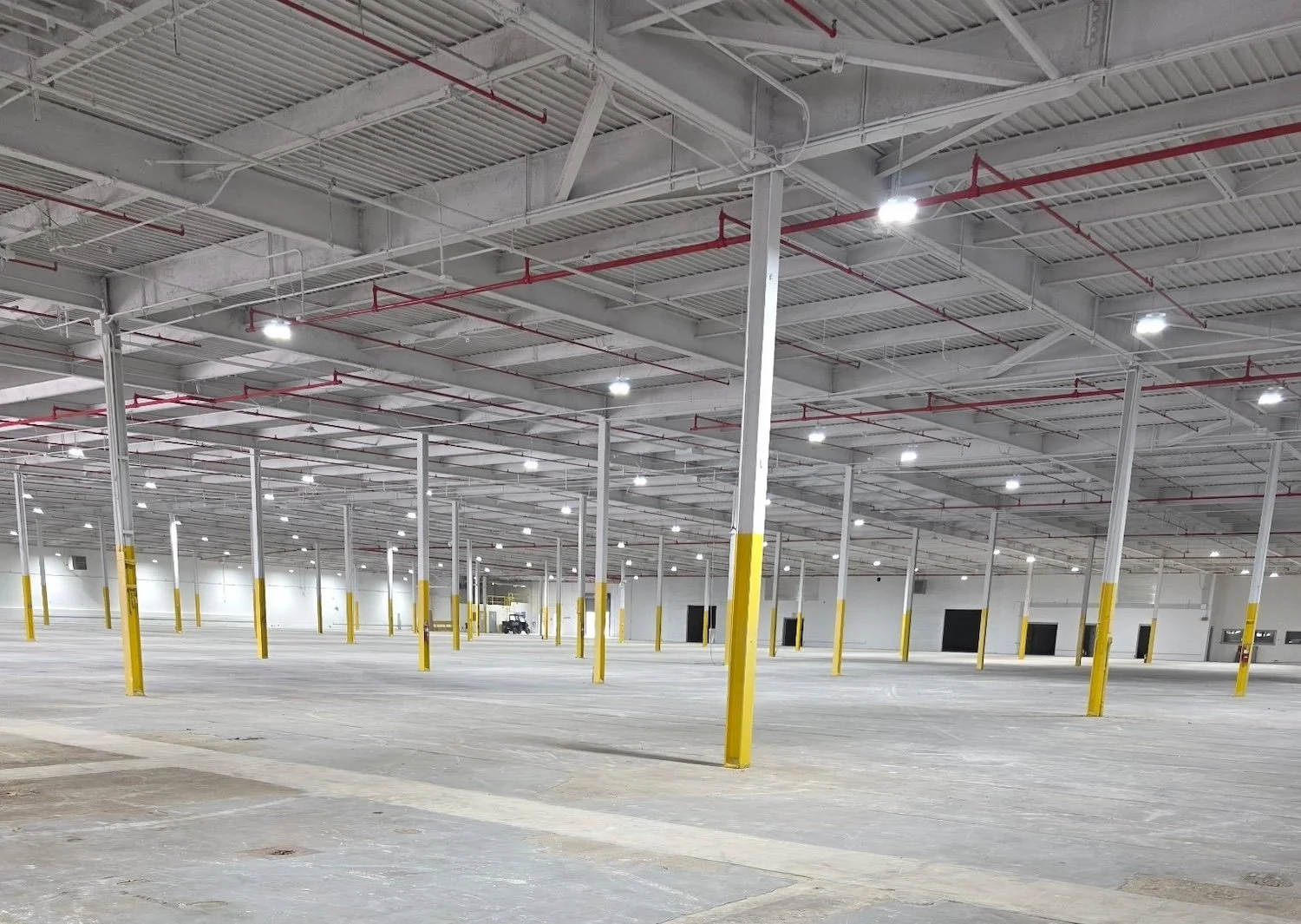 Empty indoor parking garage with concrete floors, yellow safety posts, and a high ceiling with lights and red pipes.