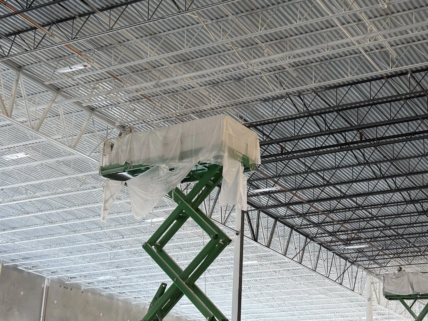 Industrial construction site with a green scissor lift elevating workers to install or repair ceiling panels covered with plastic sheets in a large building under construction.