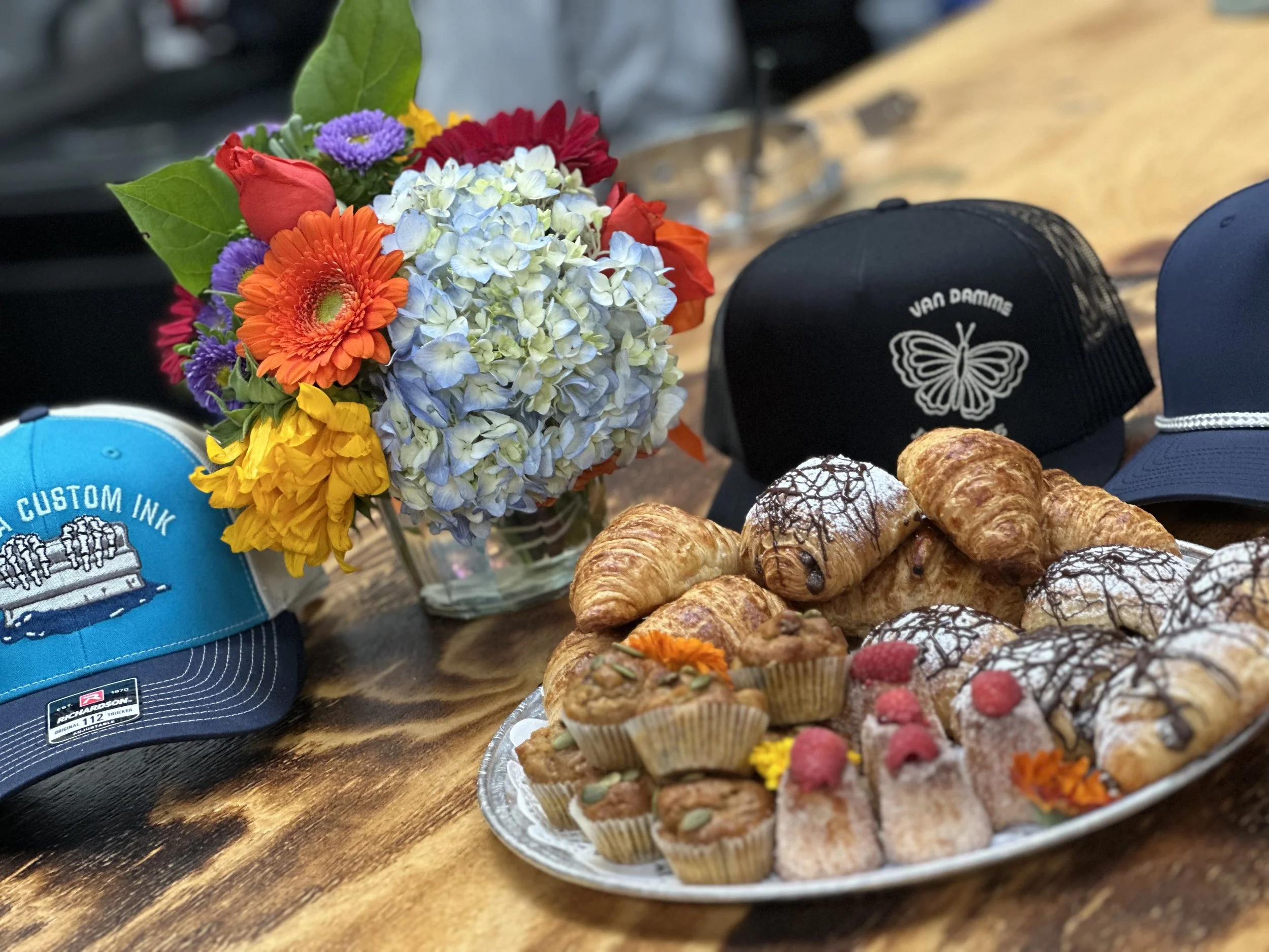 A bouquet of colorful flowers, a plate of assorted pastries including croissants and muffins, and three caps with logos and butterfly designs on a wooden table.