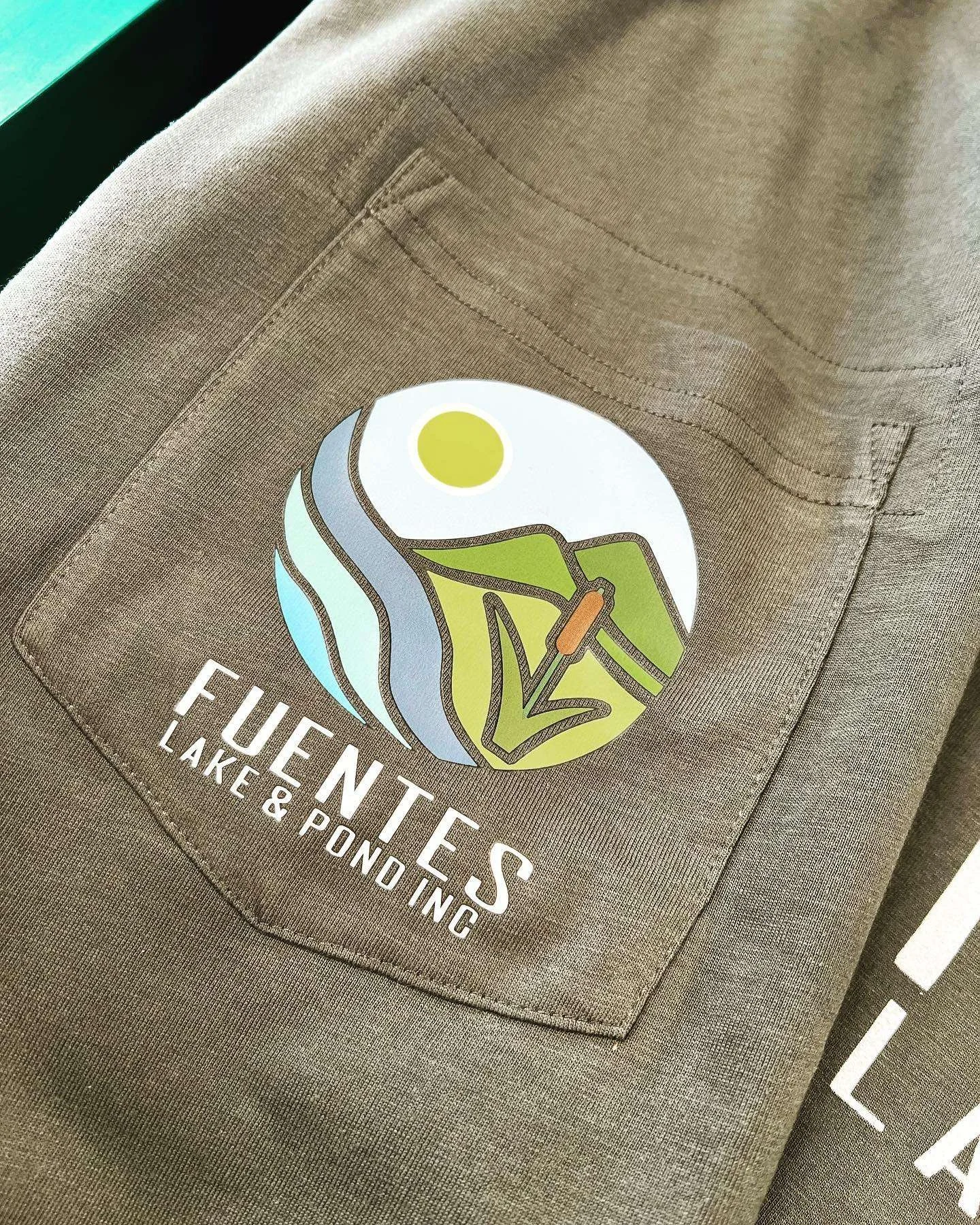 Close-up of a tan jacket pocket with a colorful logo that reads 'Fewentes Lake & Pond Inc.' The logo depicts a landscape with a sun, water, and land.