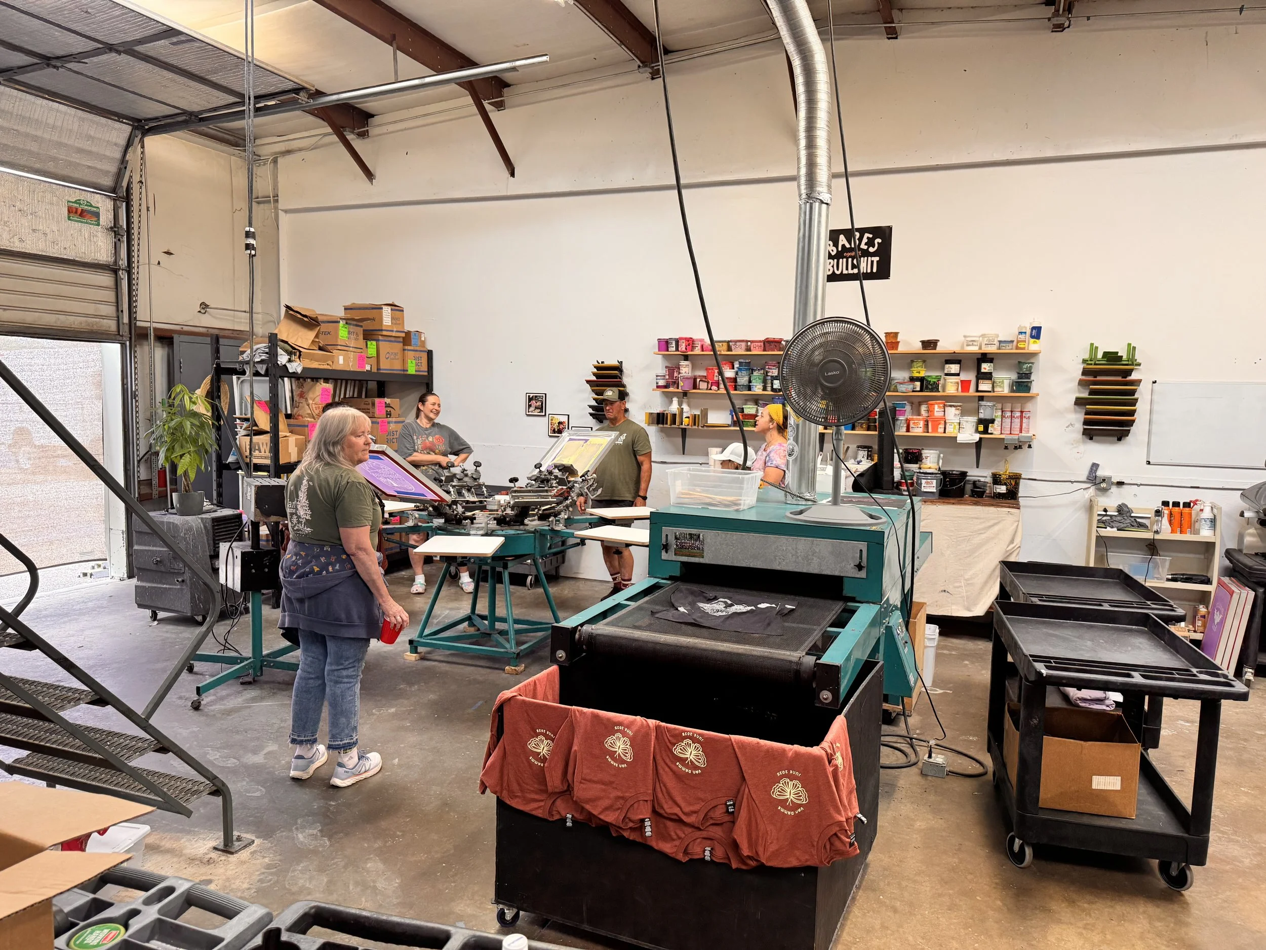 People working in a screen printing workshop, with a large printing machine, shelves with paints, and printing supplies in the background.