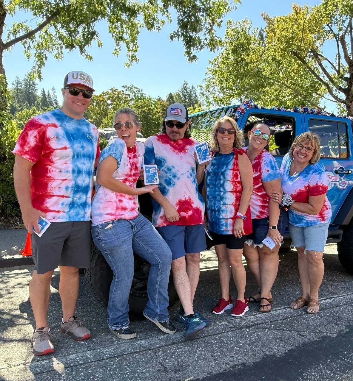 Group of six people wearing red, white, and blue tie-dye shirts, standing outdoors near a decorated vehicle, smiling and holding tickets or passes, under leafy trees on a sunny day.