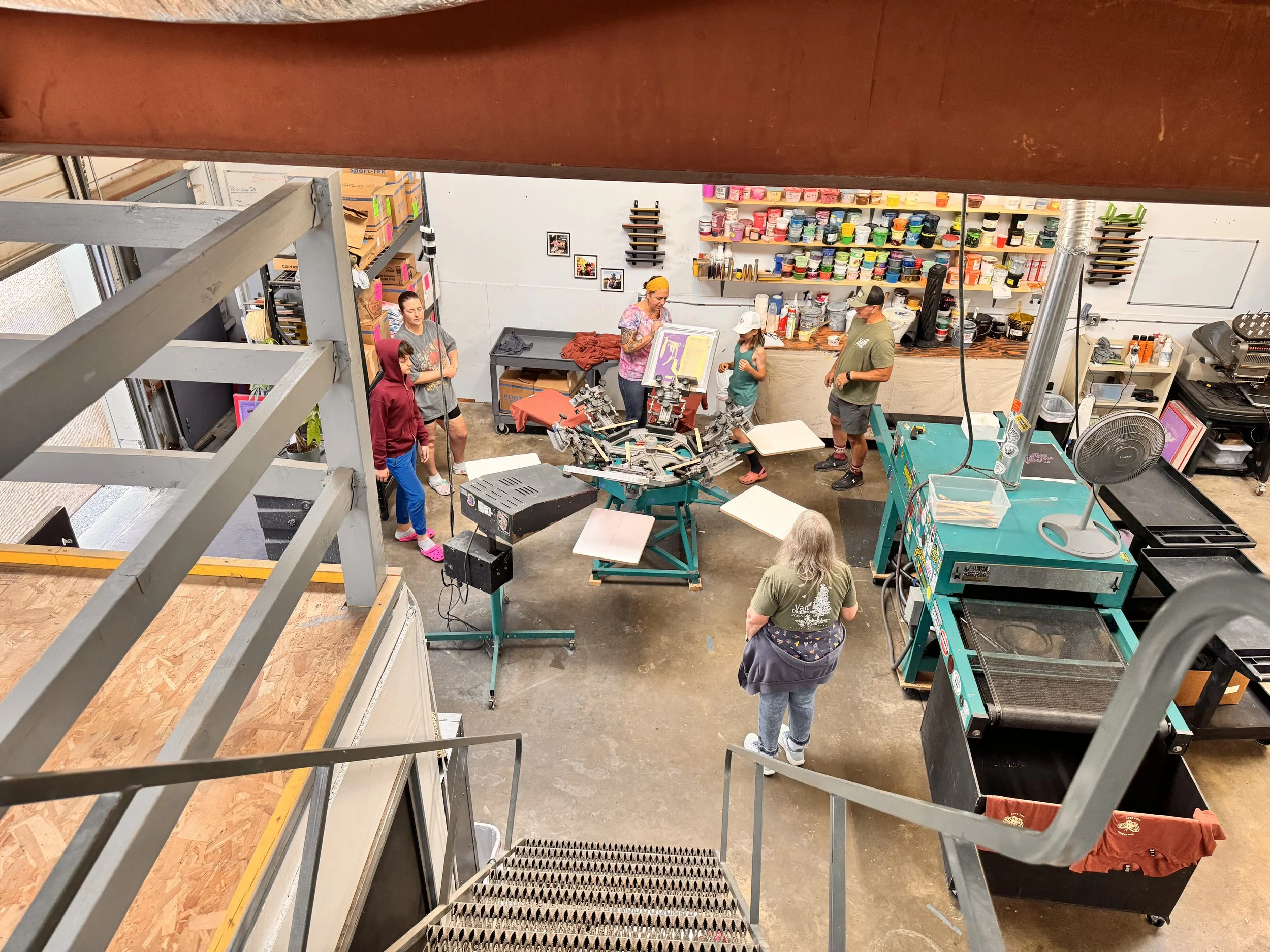 People working in a print shop or screening workshop with printing equipment, shelves of paint or ink, and printing frames.