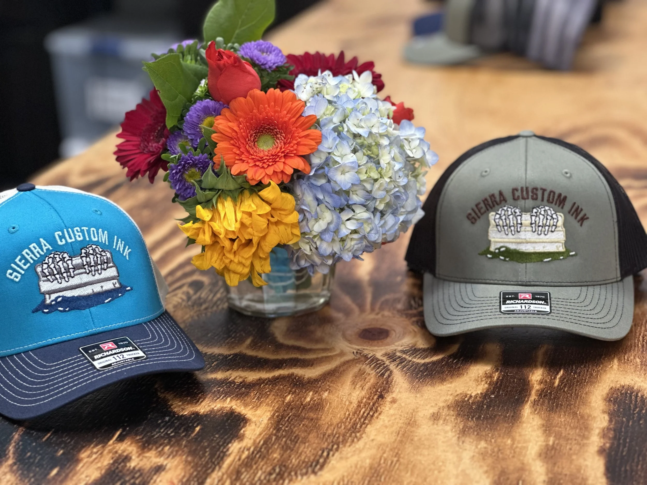 A glass vase with a colorful bouquet of flowers, flanked by two Sierra Custom Ink baseball caps on a wooden table.