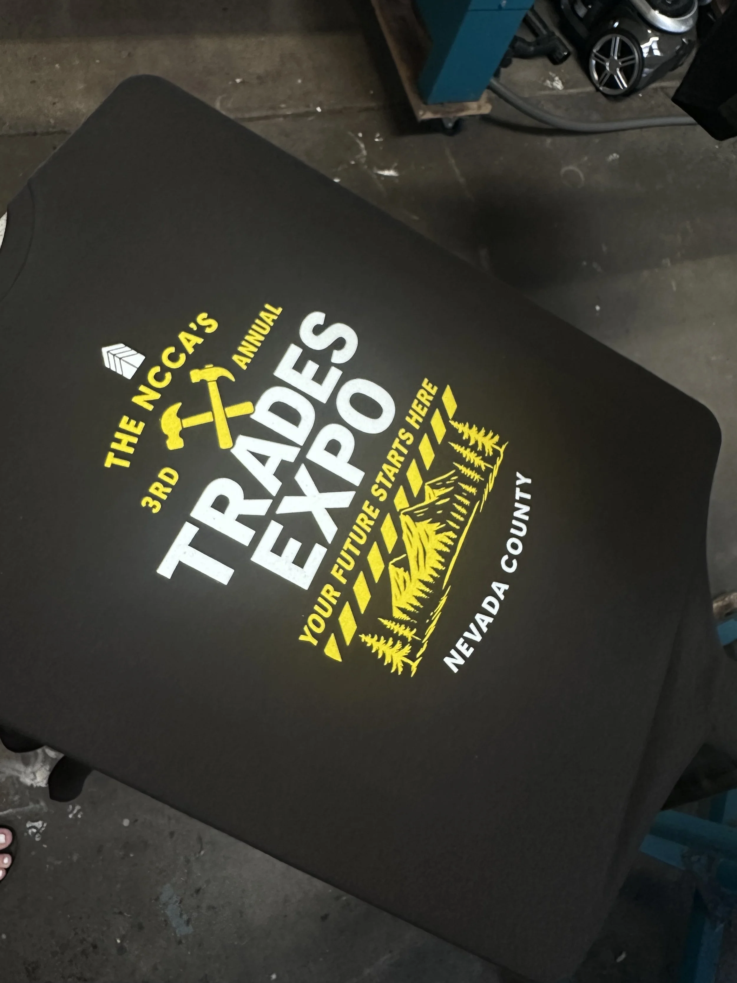A black t-shirt with yellow and white text and graphics. It reads 'The NCCA's 3rd Annual Trades Expo. Your future starts here, Nevada County' with a graphic of a hammer and crossed tools and a landscape with trees.