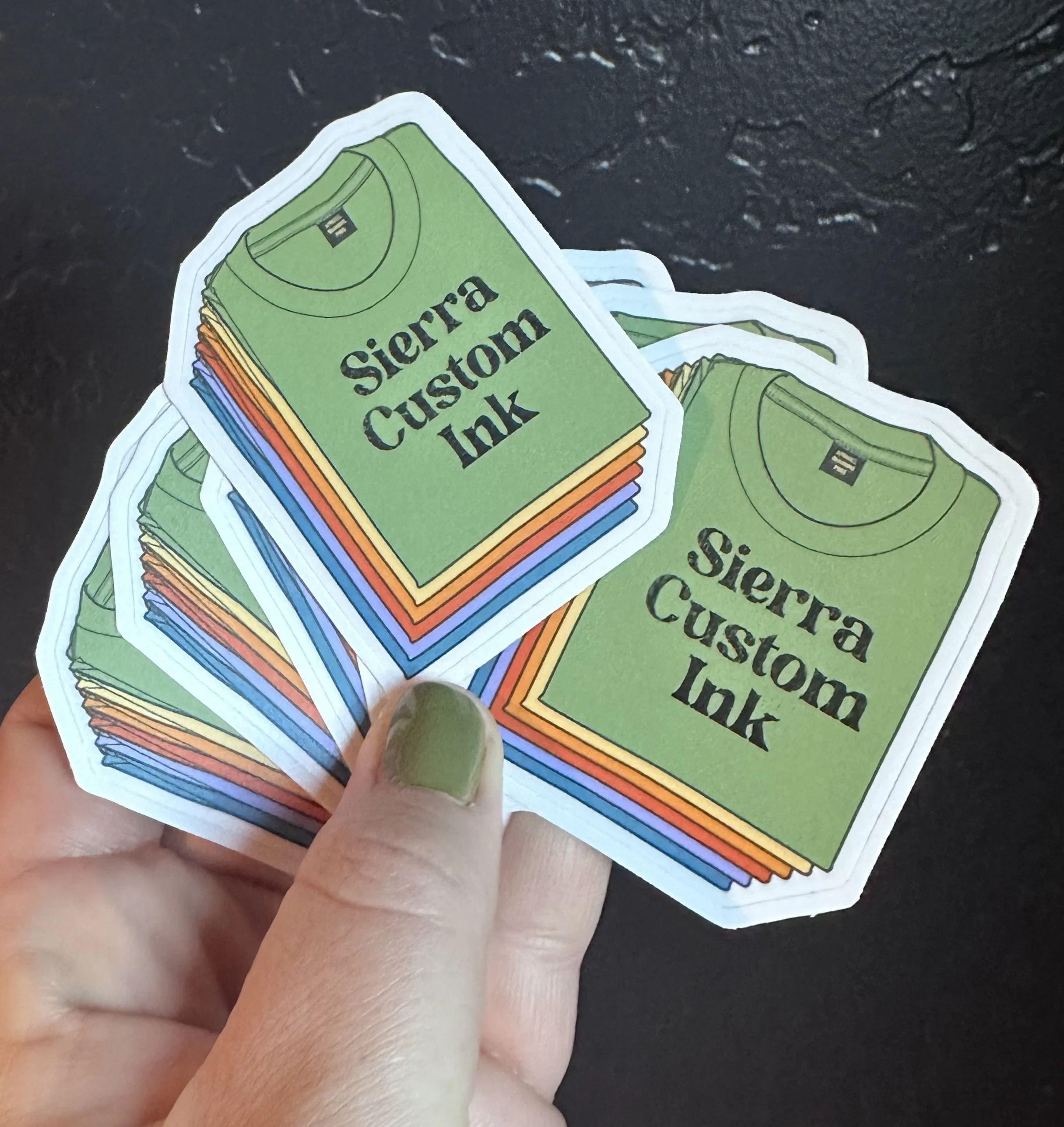 A person's hand holding a stack of colorful stickers that say 'Sierra Custom Ink' with a black shirt illustration on a green background.