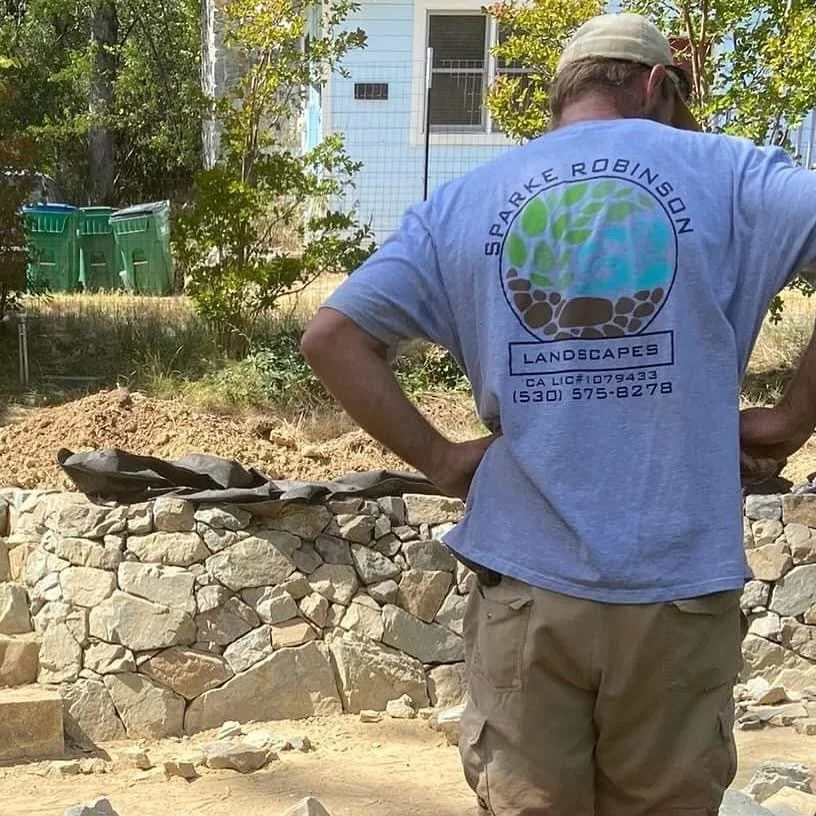Person wearing a gray T-shirt with a logo and contact information on the back, standing next to a stone wall and working on landscaping outdoors.
