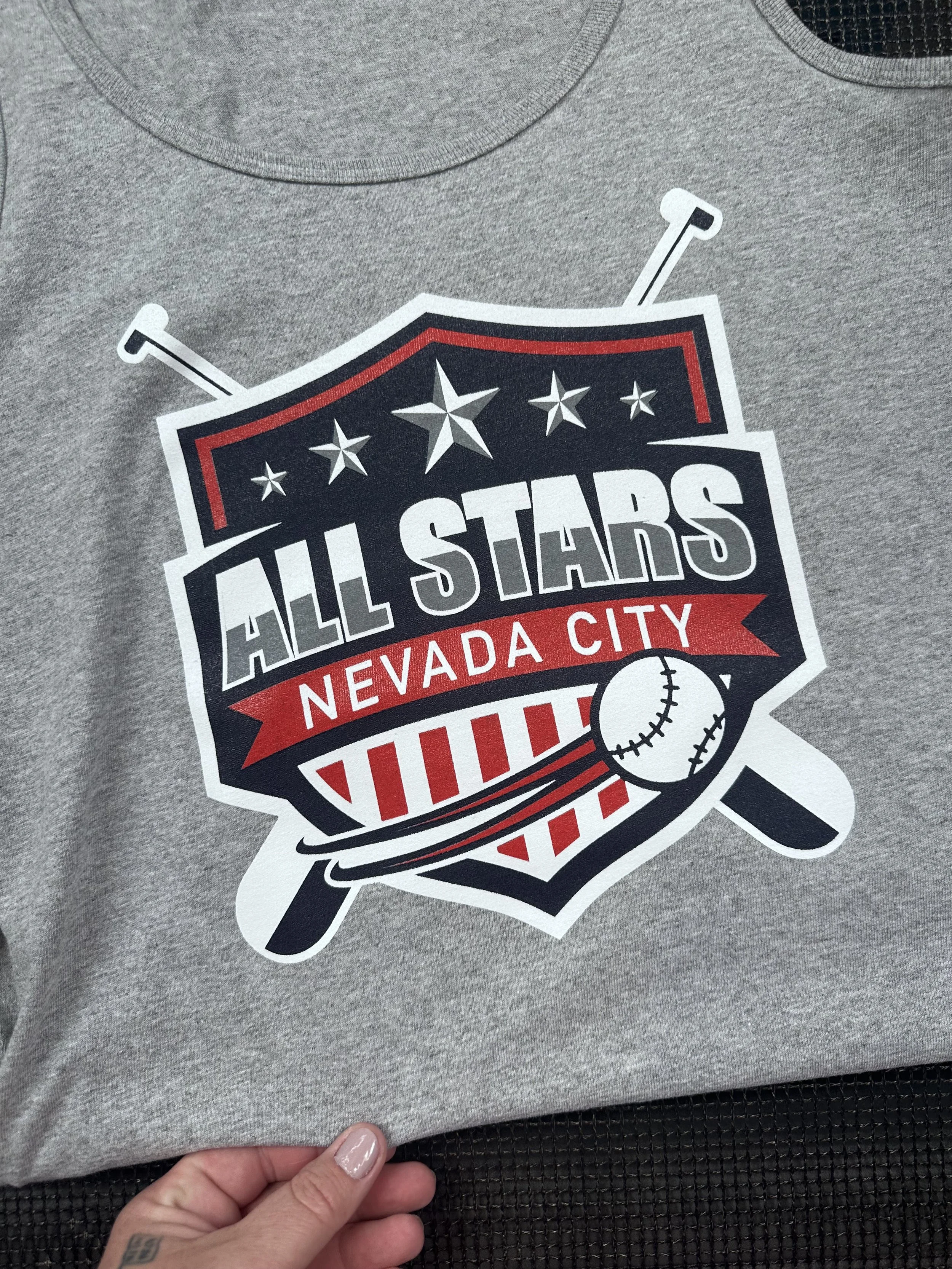 Gray t-shirt with a sports-themed design promoting Nevada City All Stars, featuring stars, baseball references, and the words Nevada City.