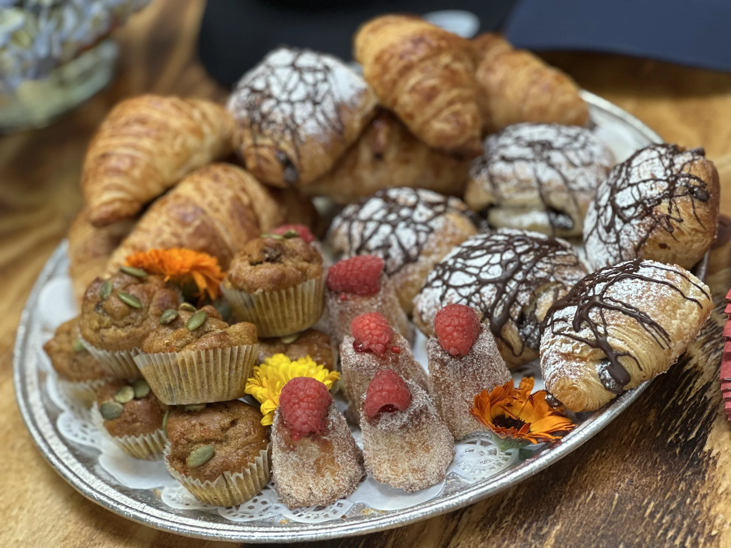 Assorted pastries and cupcakes on a decorative silver platter, including croissants, puff pastries with chocolate drizzle, muffins with fruit and pumpkin seeds, and small sugar-coated candies topped with raspberries, garnished with colorful edible fl