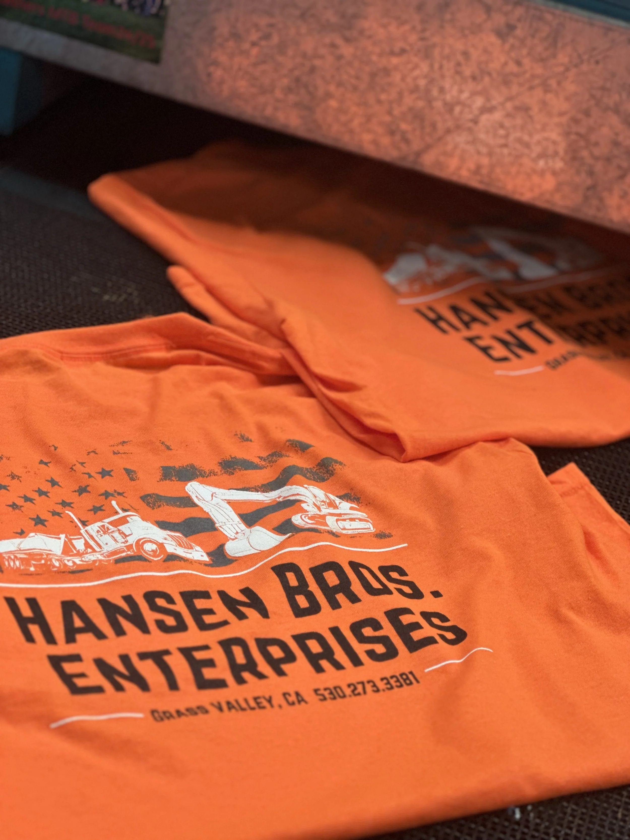 Orange T-shirts with black text and graphics for Hansen Bros. Enterprise, Inc., featuring a graphic of construction equipment and stars, displayed on a black surface.
