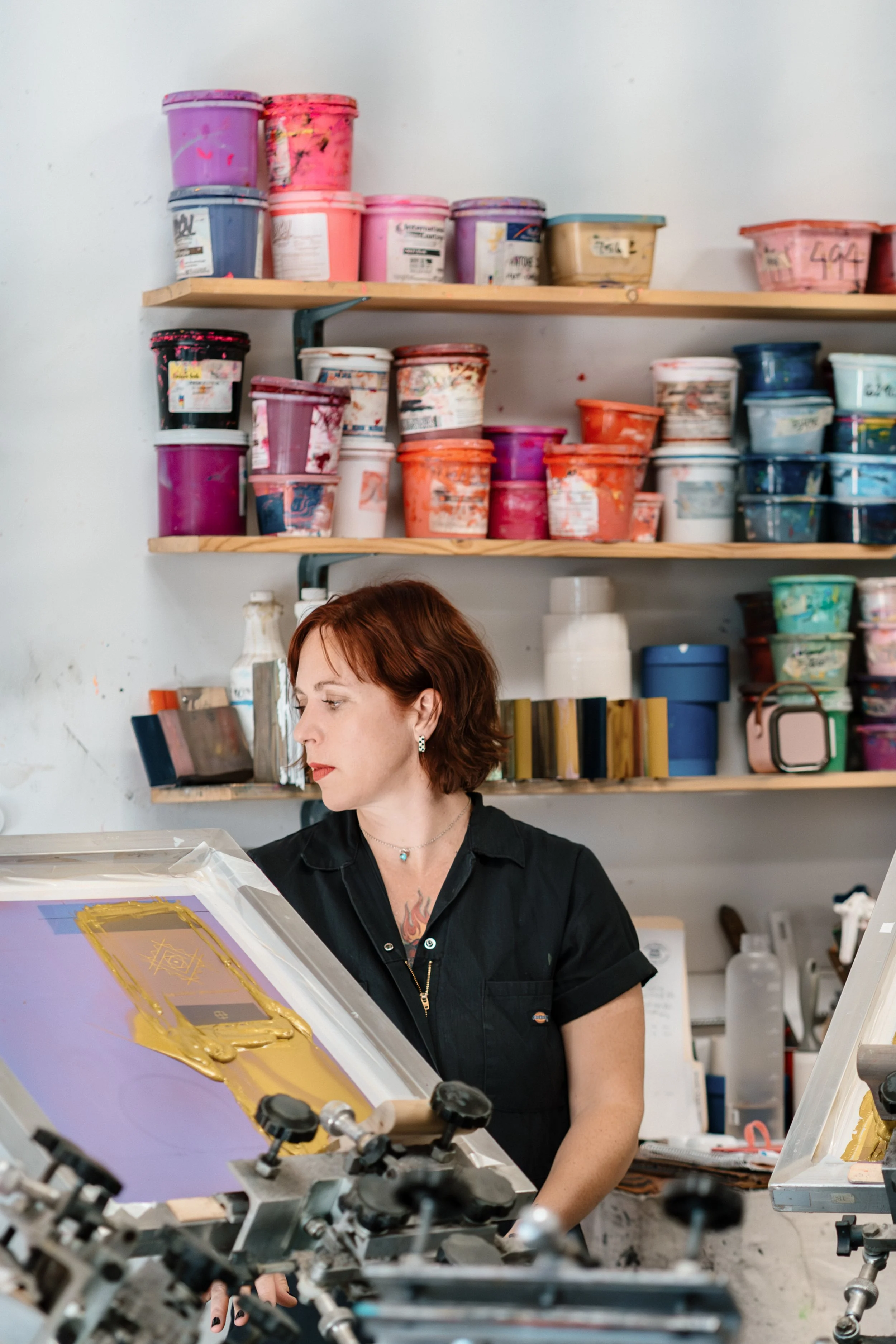 A woman with short auburn hair, wearing a black short-sleeve shirt, working in a print shop or art studio. She is focused on a screen printing press with a golden frame design. Behind her, shelves are filled with colorful containers of ink and other art supplies.