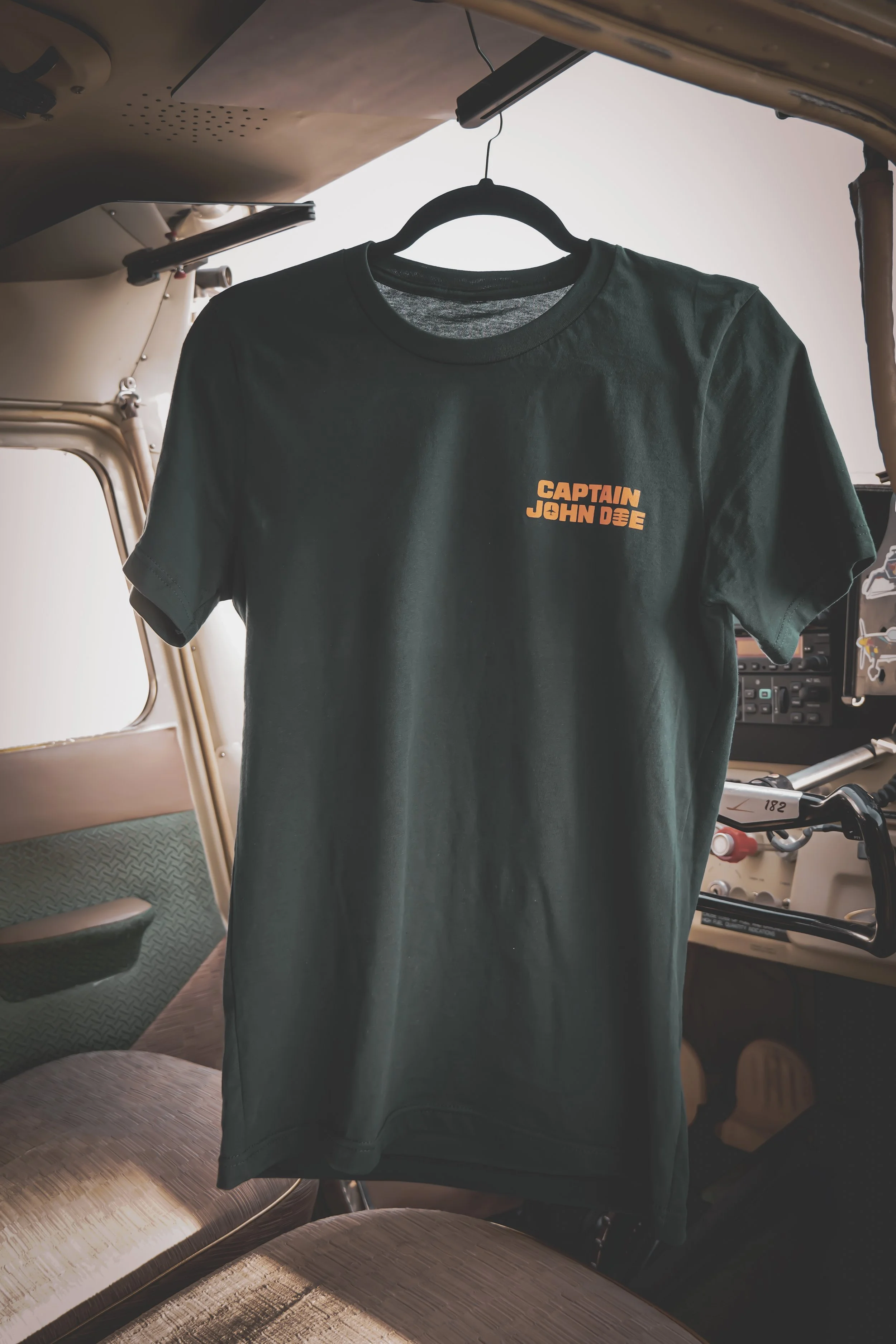 A black T-shirt hanging on a hanger inside a small aircraft cockpit. The shirt has orange text that reads, 'CAPTAIN JOHN DOE'.