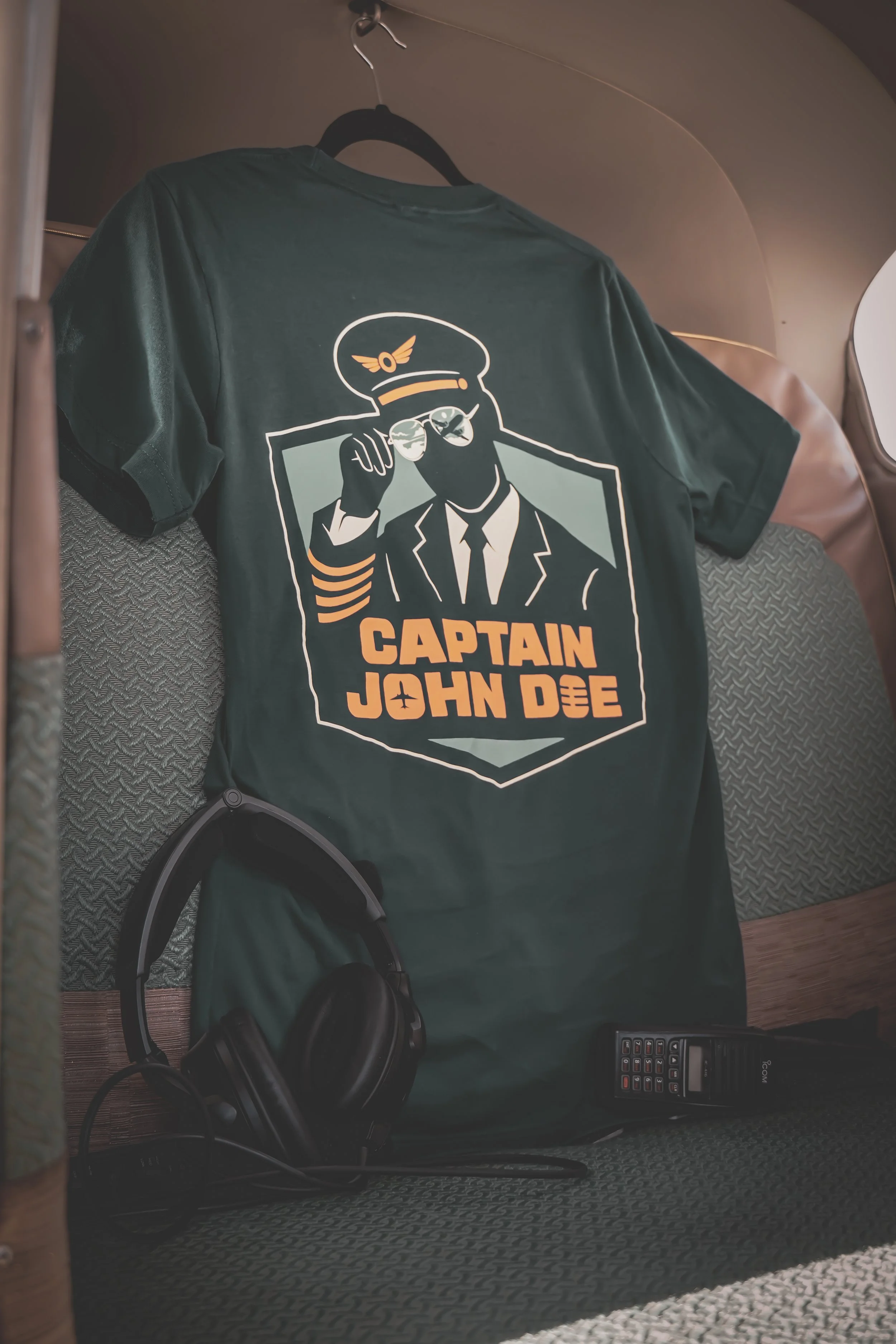 A green t-shirt with a graphic of a pilot wearing glasses and a captain's hat, with the text "Captain John Doe" on it. The t-shirt hangs on a hanger in a plane, and there are black headphones and a radio placed on the seat below the t-shirt.
