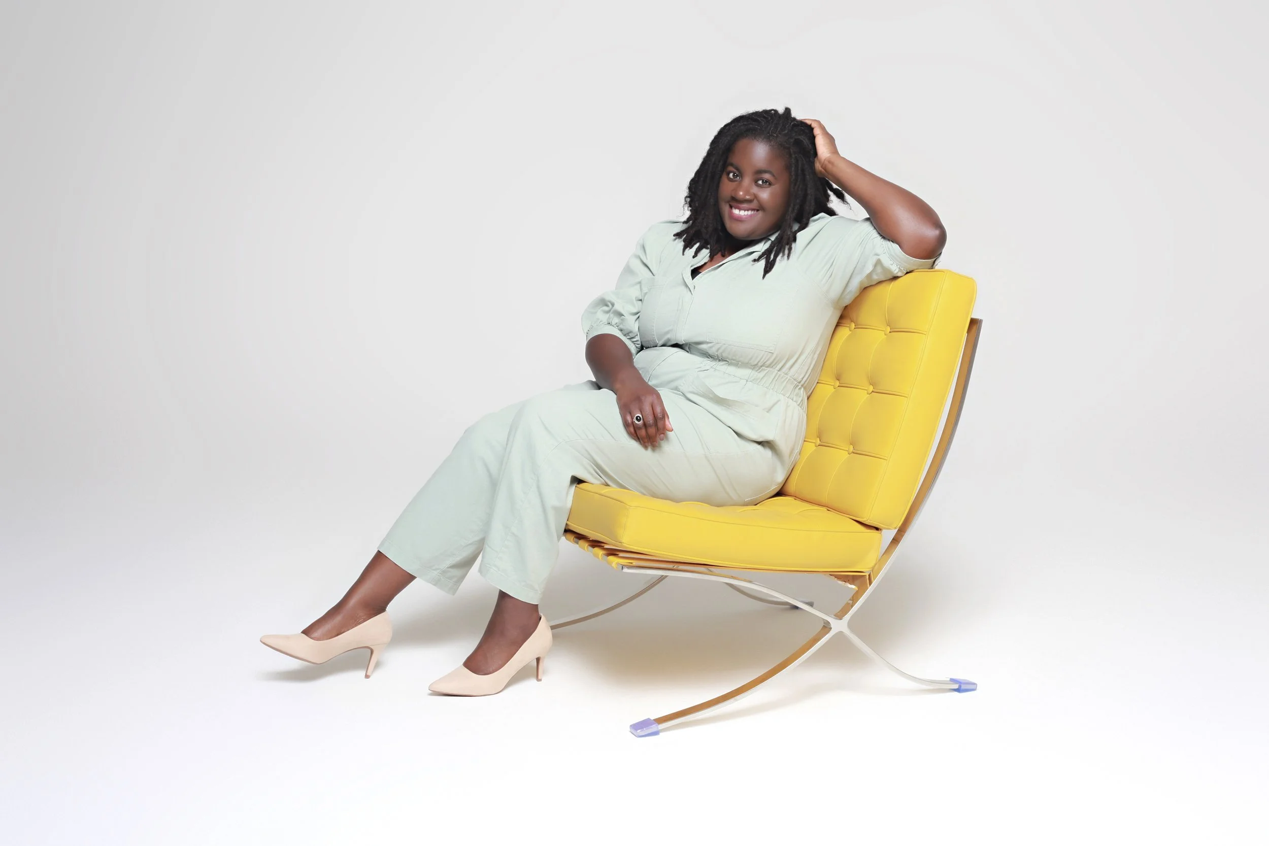 Smiling woman sitting on a yellow lounge chair against a plain white background, wearing a light-colored outfit and beige high heels.